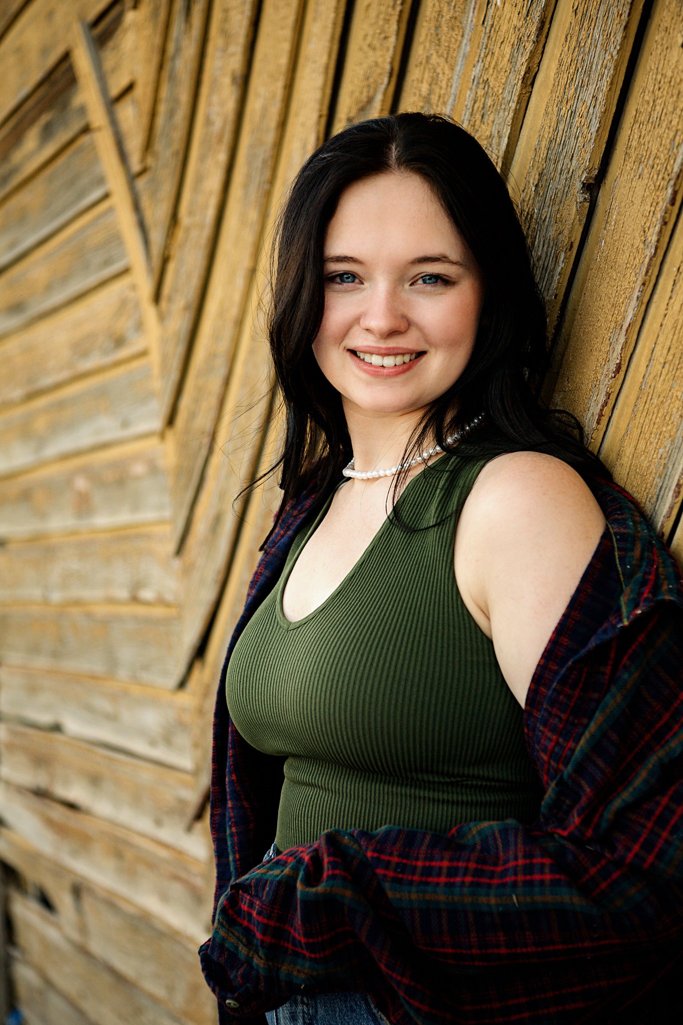 A young woman with long dark hair smiling, wearing a green ribbed tank top, a pearl necklace, and a red plaid shirt, leaning against a wooden wall.