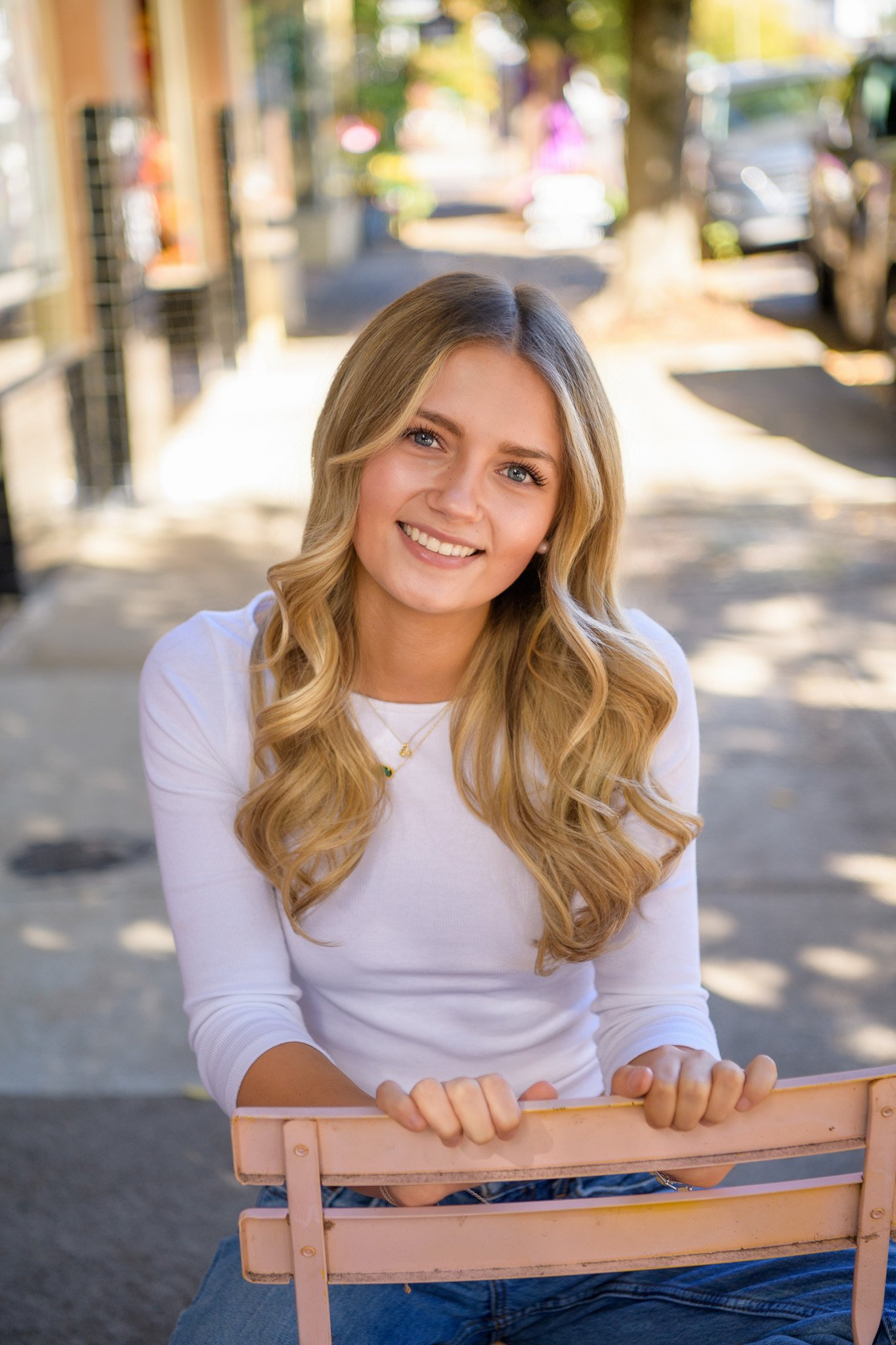 A young woman with long blonde hair and blue eyes sitting outdoors on a pink bench in a sunny, urban setting, smiling at the camera.