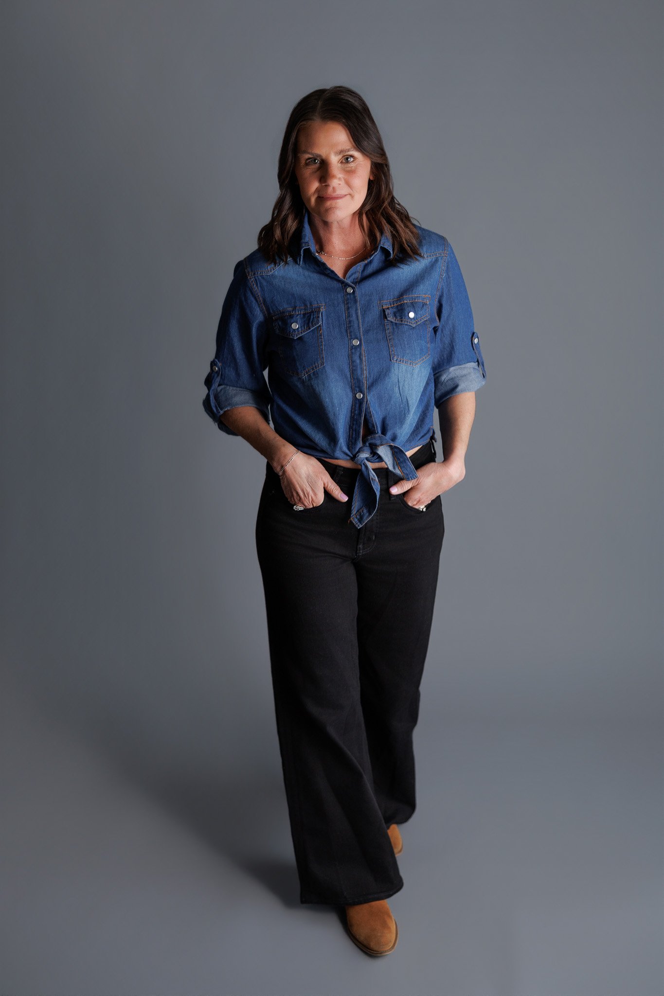 A woman with shoulder-length brown hair, wearing a denim shirt tied at the waist, black pants, and tan boots, stands against a gray background.