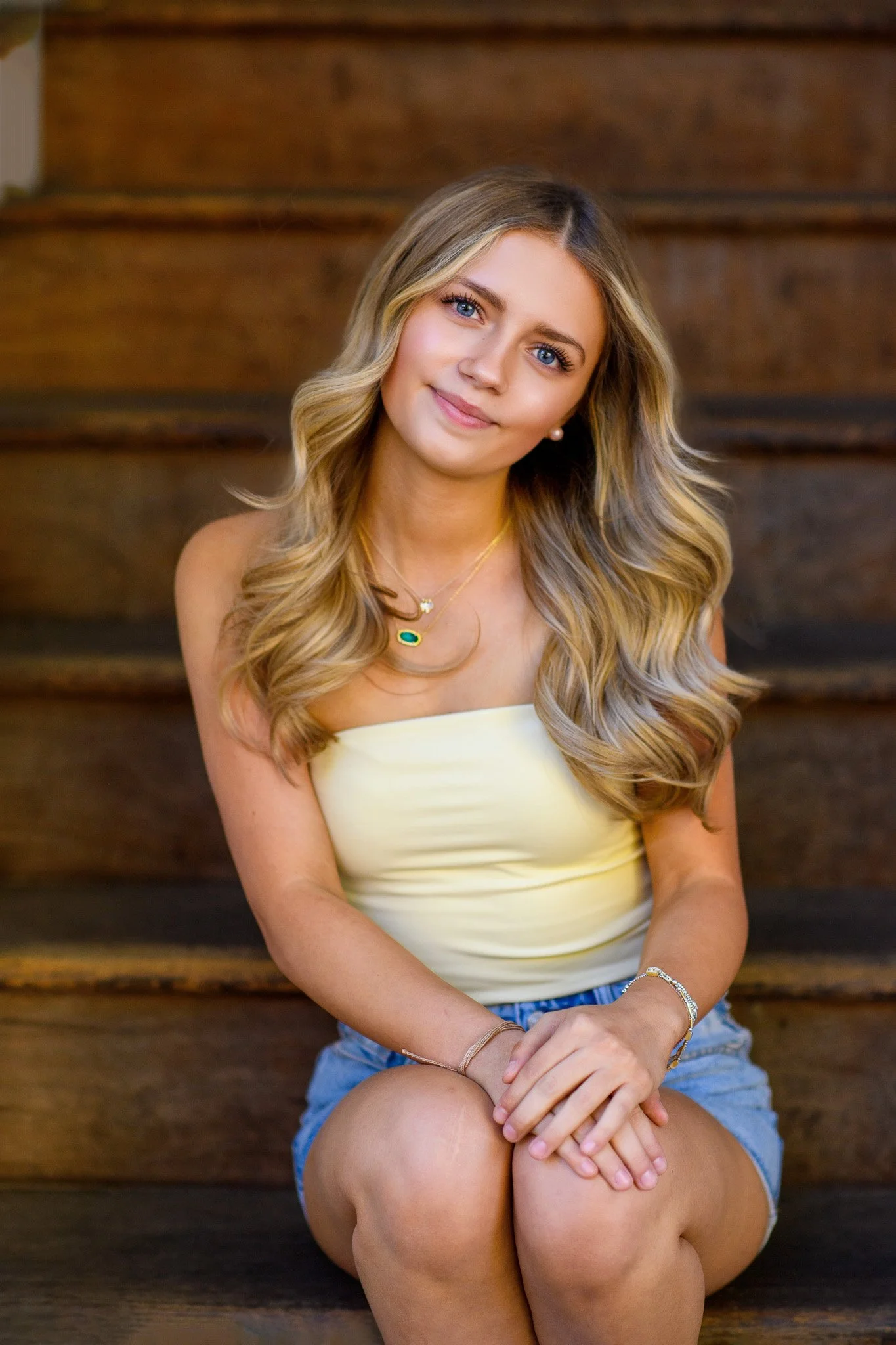 A young woman with long, wavy blonde hair, dressed in a yellow top and denim shorts, sitting on wooden stairs, smiling at the camera.