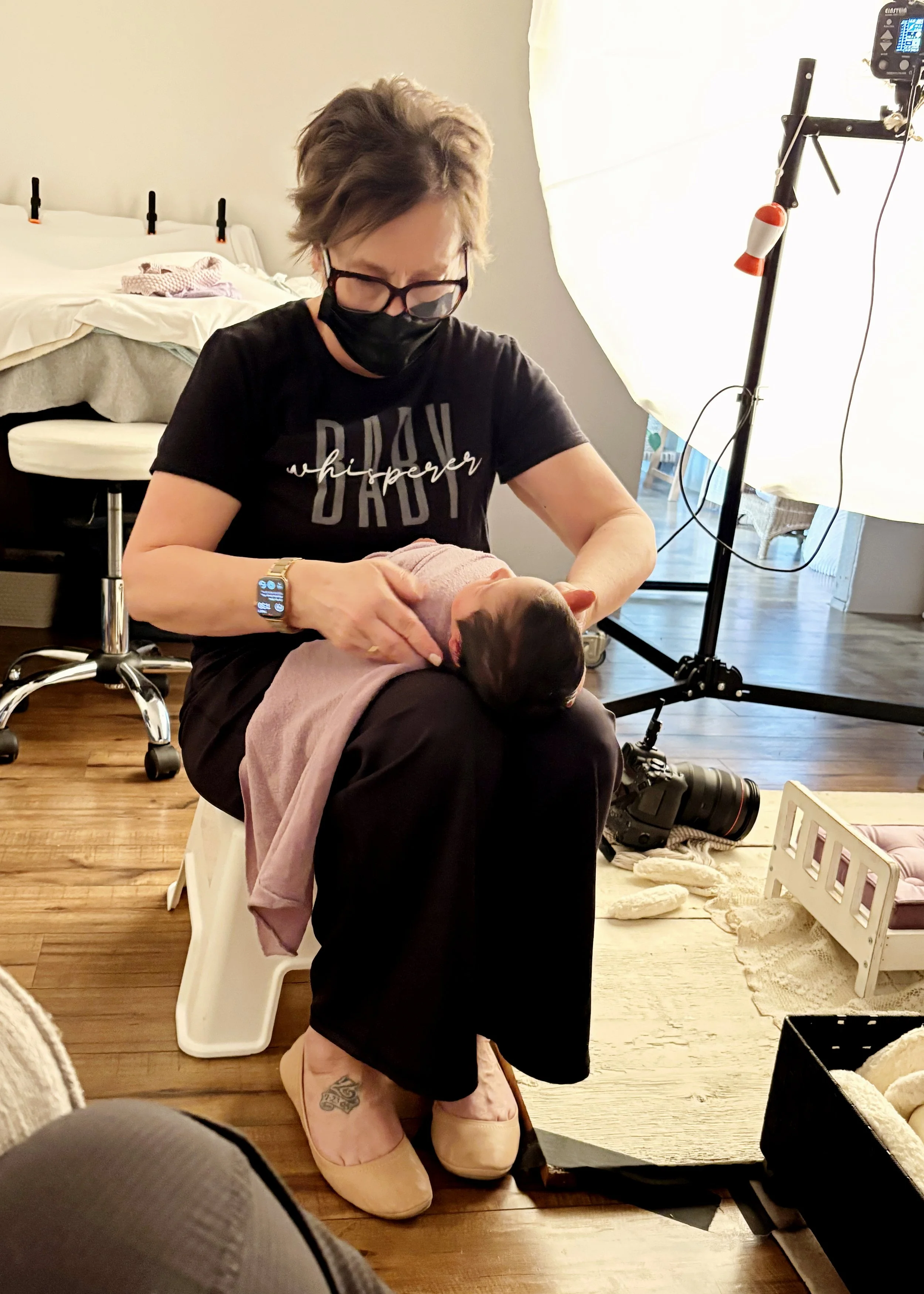 A woman wearing a black face mask, glasses, and a black t-shirt that says ‘baby whisperer’ is sitting on a white step stool, breastfeeding a newborn baby wrapped in a pink blanket. She is indoors with a camera and lighting equipment nearby, and a bed in the background.