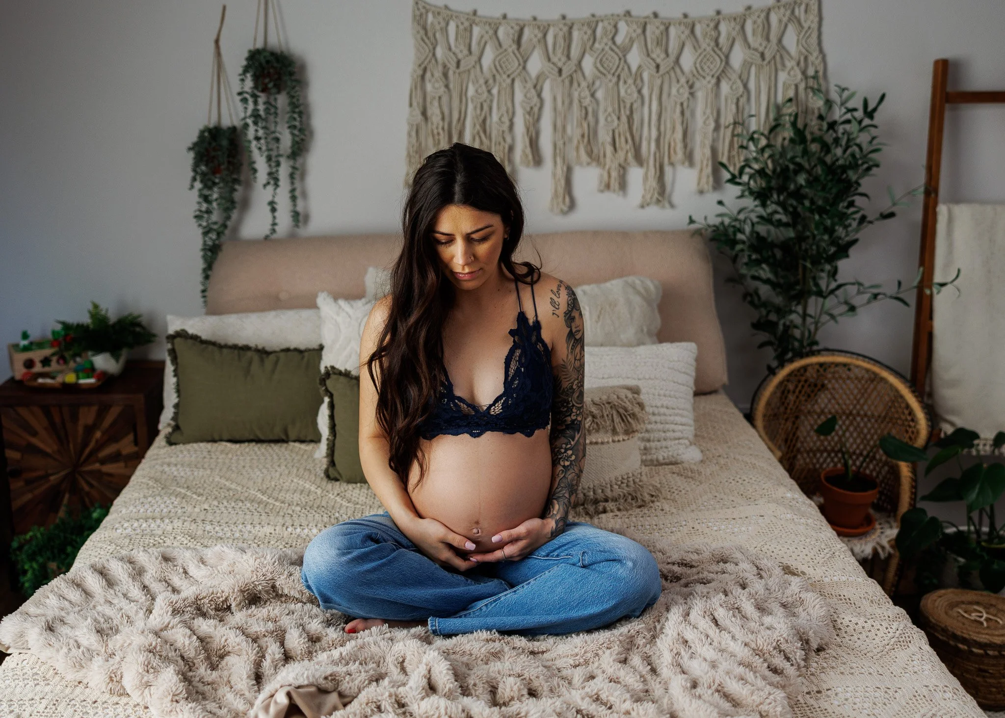 Pregnant woman sitting cross-legged on a bed, holding her belly and looking down, in a cozy bedroom with plants and boho decor.