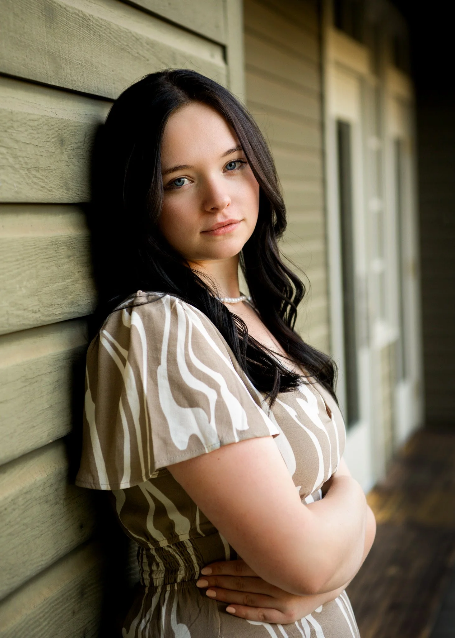 A young woman with long dark hair and blue eyes, wearing a beige patterned dress and pearl necklace, leaning against a green wooden wall with arms crossed.