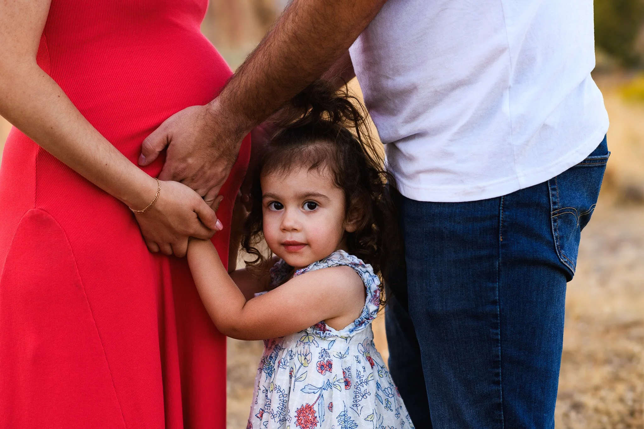 A young girl hugging her pregnant mother, with her father nearby, outdoors.