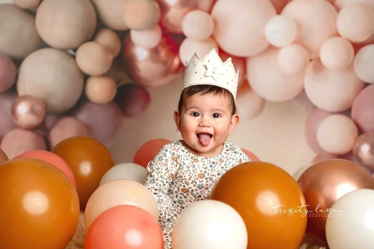 A 1st birthday baby girl with short hair, wearing a crown hat and a floral dress, sitting among metallic and pastel-colored balloons, sticking out her tongue and smiling with an open mouth.