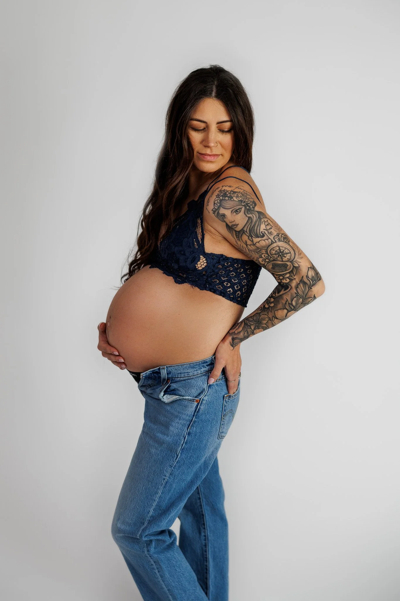 Pregnant woman with long brown hair wearing a dark blue lacy crop top and blue jeans, standing against a plain white background.