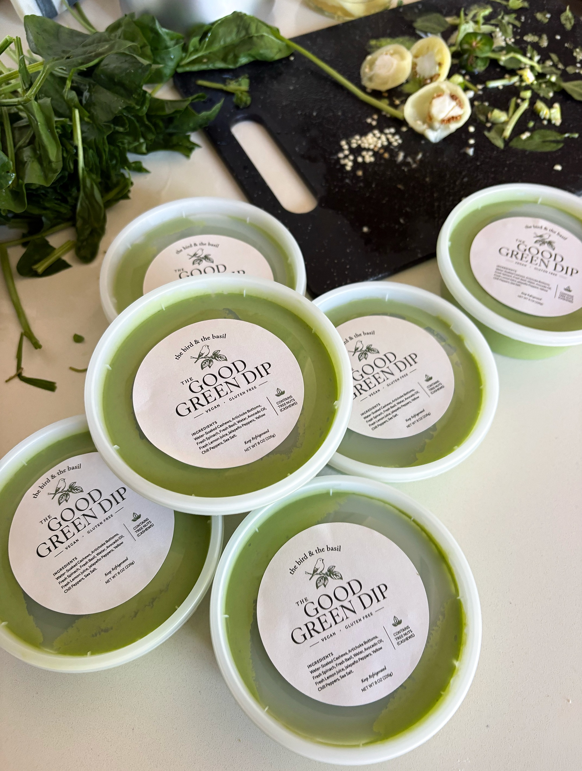 The bird & the basil The Good Green Dip labels on packages of dip on kitchen counter with fresh ingredients in the background