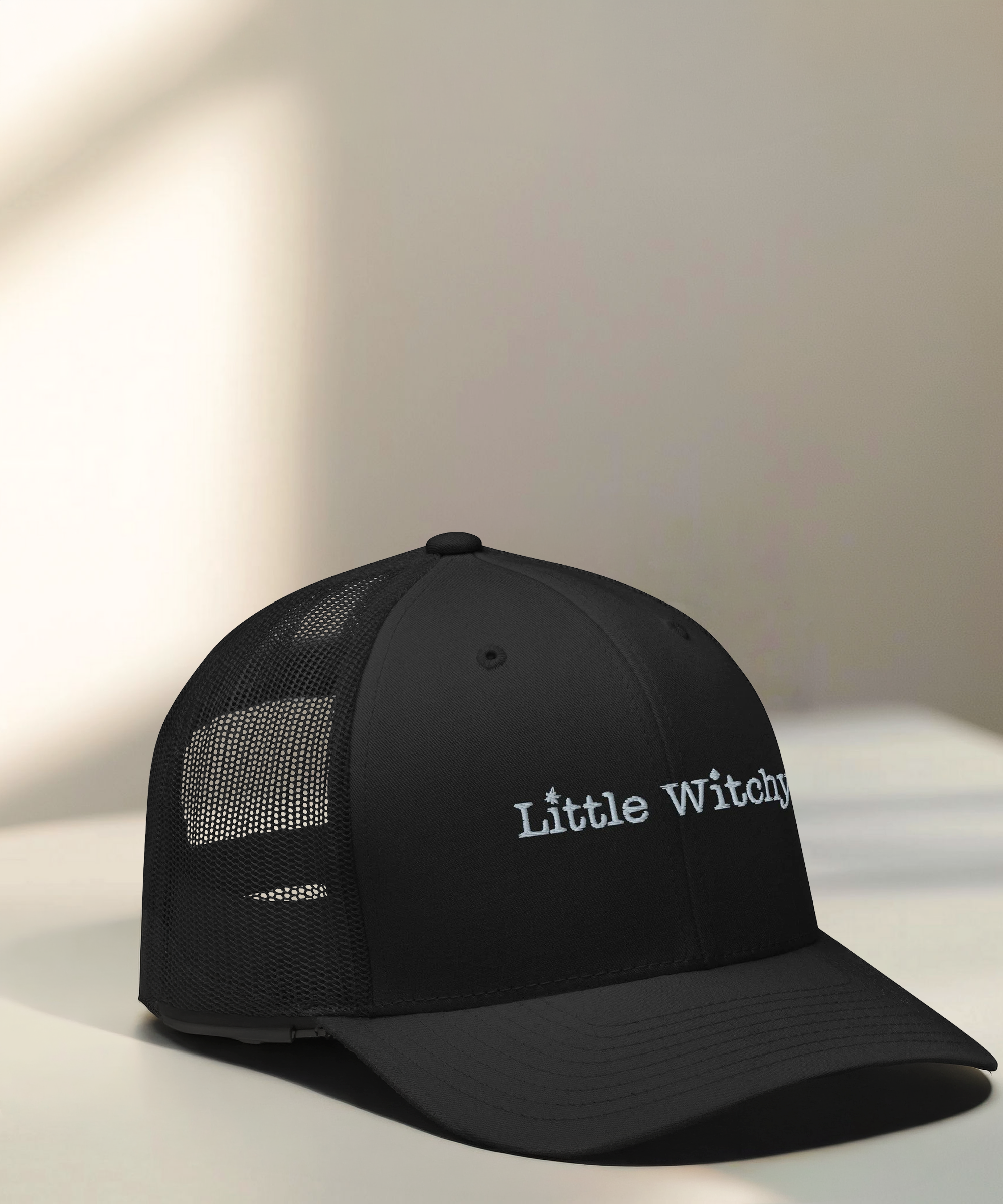 Little Witchy Trucker Hat angled view on a light colored beige table with sunlight and shadows