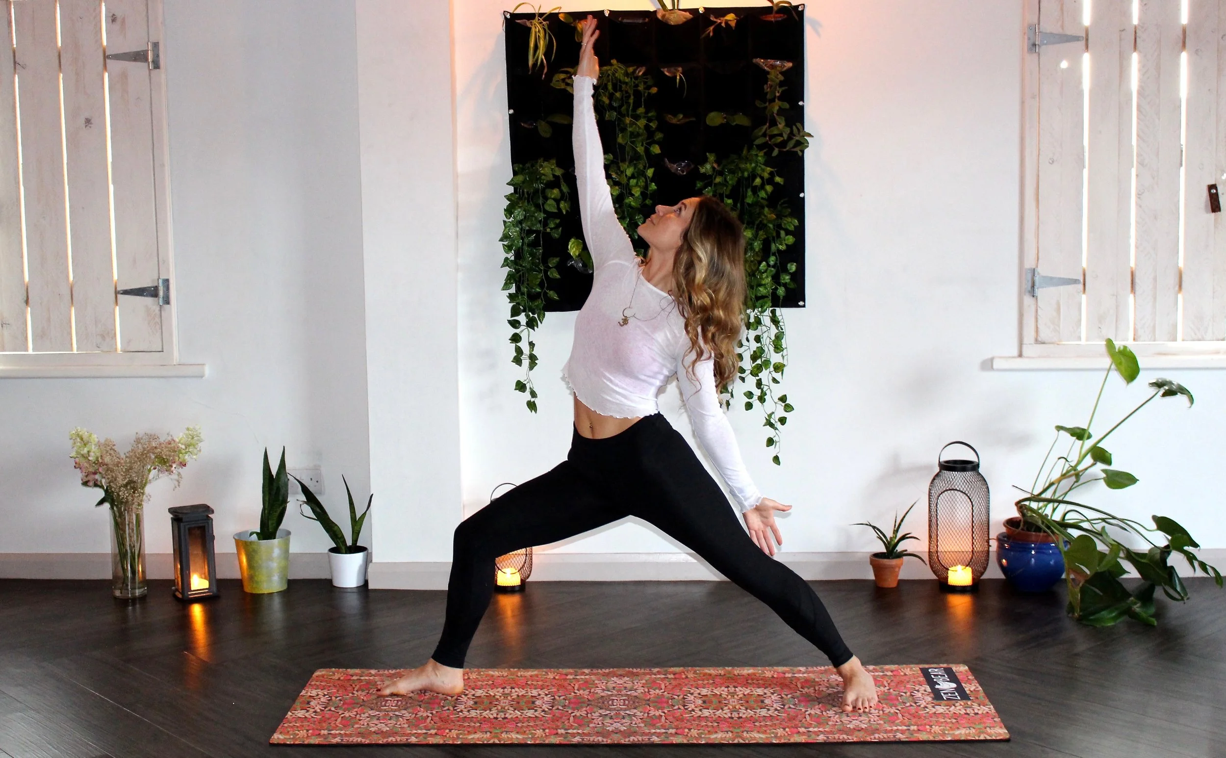 Photo by Zen Bear Yoga on Unsplash A woman is standing in warrior 2 pose wearing a long-sleeved white shirt and long black yoga pants. She is on a mat that looks like a persian carpet in a warm white room with candles.