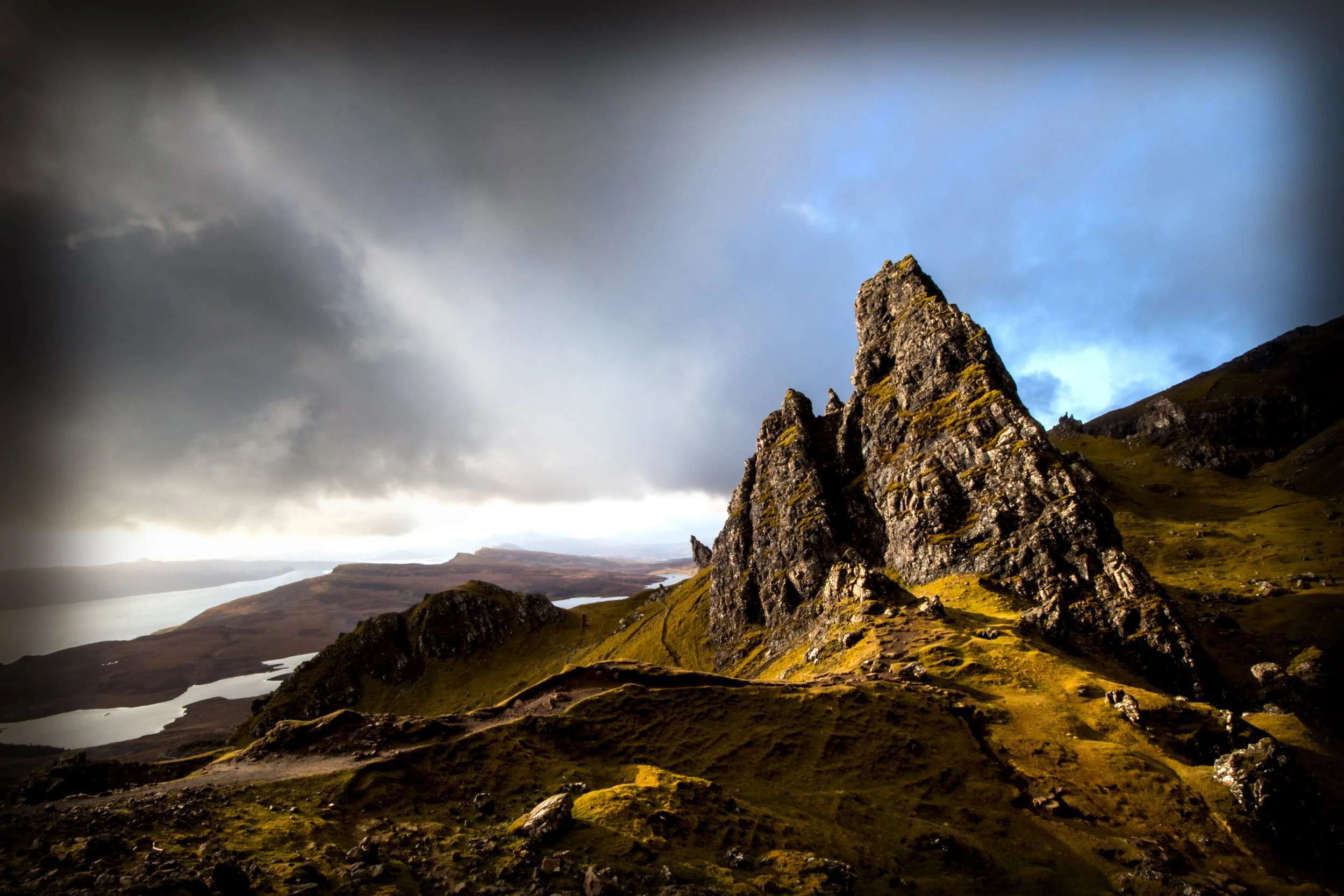 A rocky mountain with steep slopes, surrounded by grassy hills and lakes, under a cloudy sky with patches of blue.