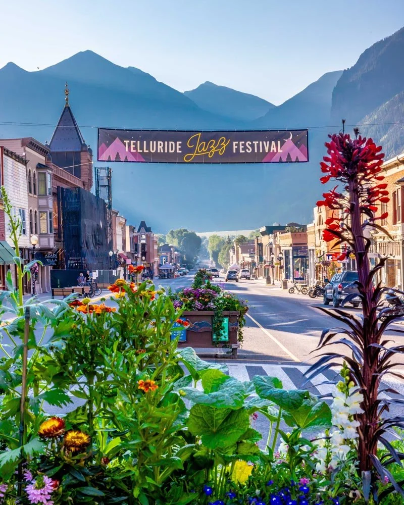 **Alt text:** Telluride's historic Main Street decorated with a Telluride Jazz Festival banner suspended overhead, welcoming visitors to the 2026 festival with the San Juan Mountains in the background.