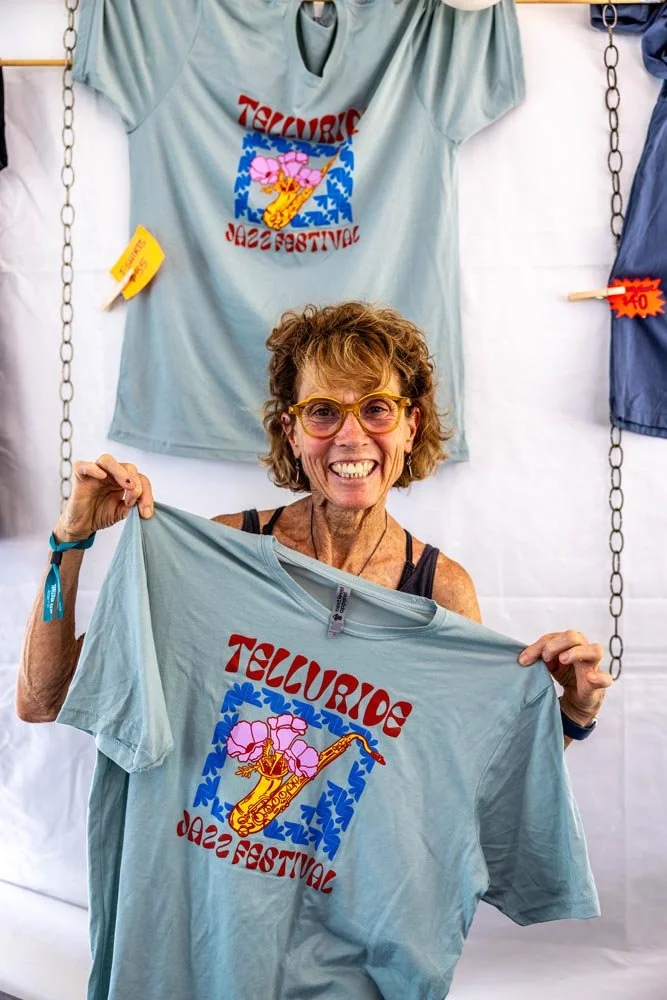 **Alt text:** Volunteer proudly holds up official Telluride Jazz Festival merchandise inside the festival merch tent, showcasing apparel and souvenirs available during the 2026 festival in Telluride Town Park.
