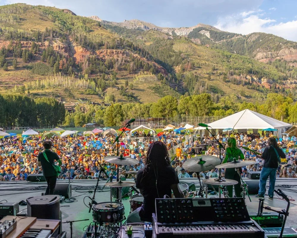 Musicians perform live on the main stage during the 2026 Telluride Jazz Festival, entertaining the crowd in Telluride Town Park with the scenic San Juan Mountains as a backdrop.