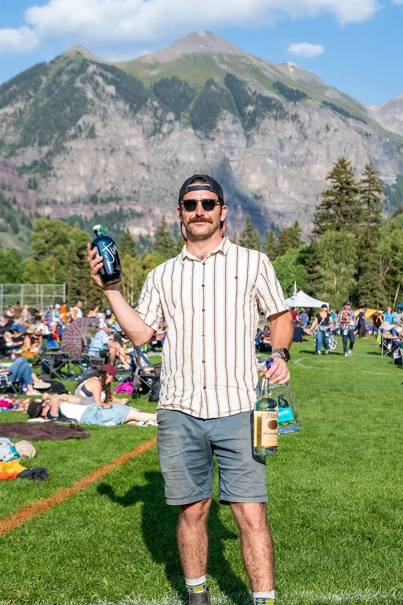 Certified Sommelier and Festival Mixologist Andrés Vargas-Johnson poses with premium spirits against the scenic San Juan Mountains, showcasing the craft cocktail program for the Society Club experience at the 2026 Telluride Jazz Festival.