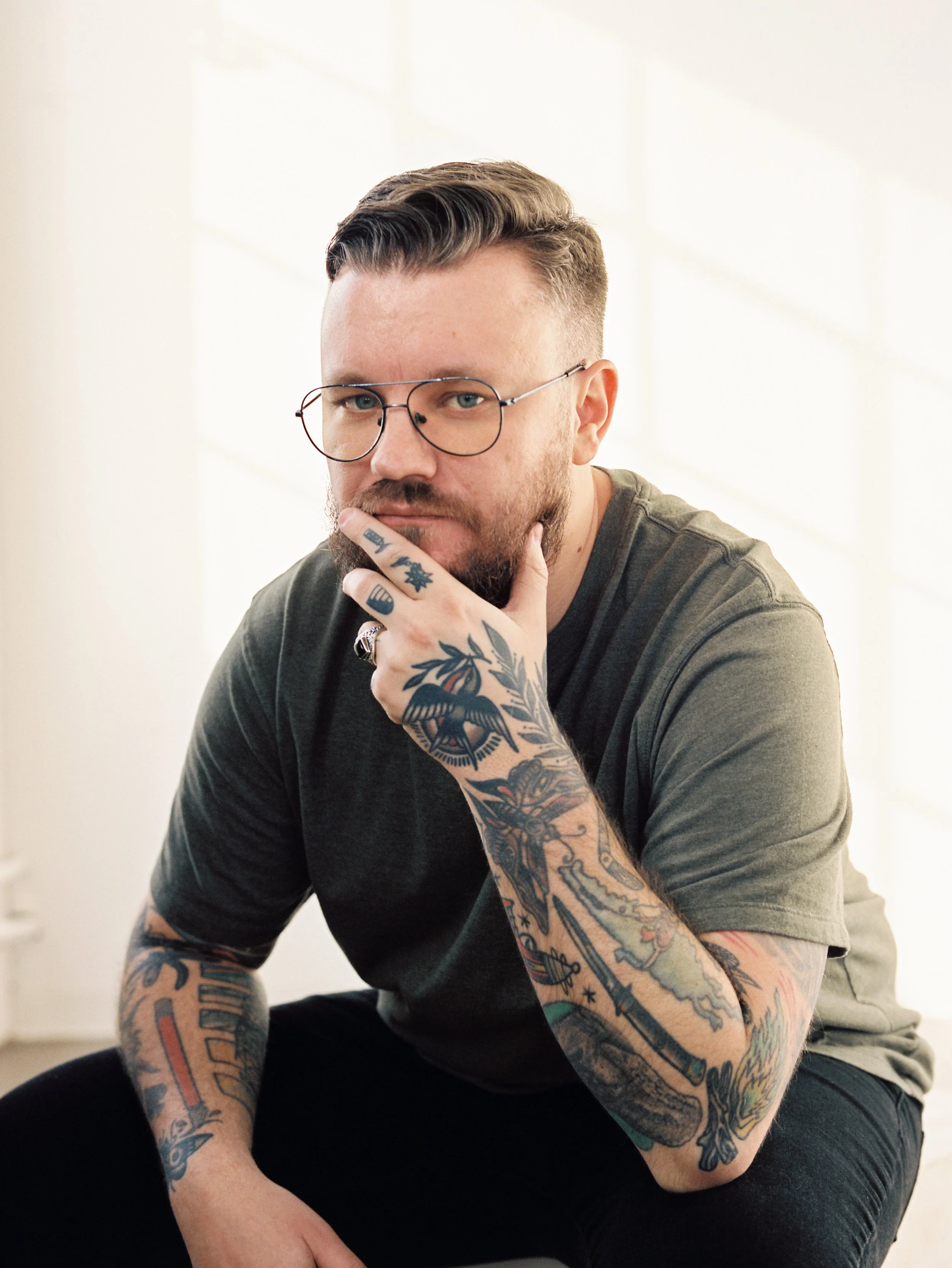 A man with glasses, tattoos on arms, beard, and styled hair, looking directly at the camera with hand on chin, in a bright room with natural light.