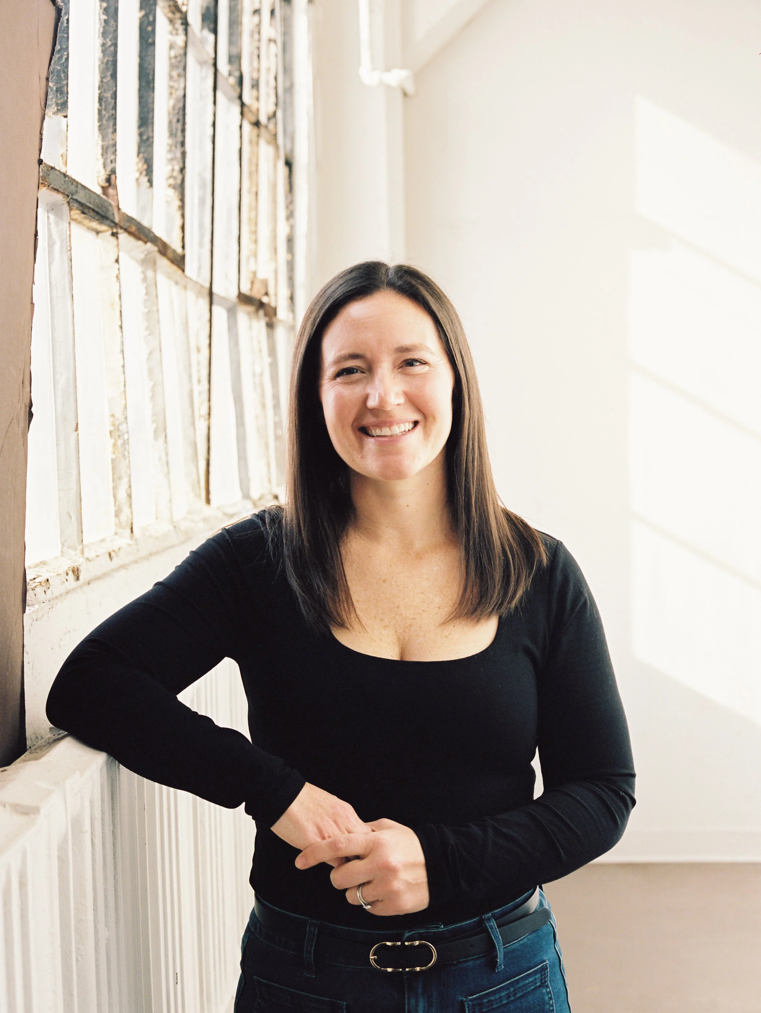 Portrait of a woman with shoulder-length dark brown hair, smiling, wearing a black long-sleeve shirt and jeans, standing indoors near a wall with peeling paint and glass blocks, with sunlight coming through.