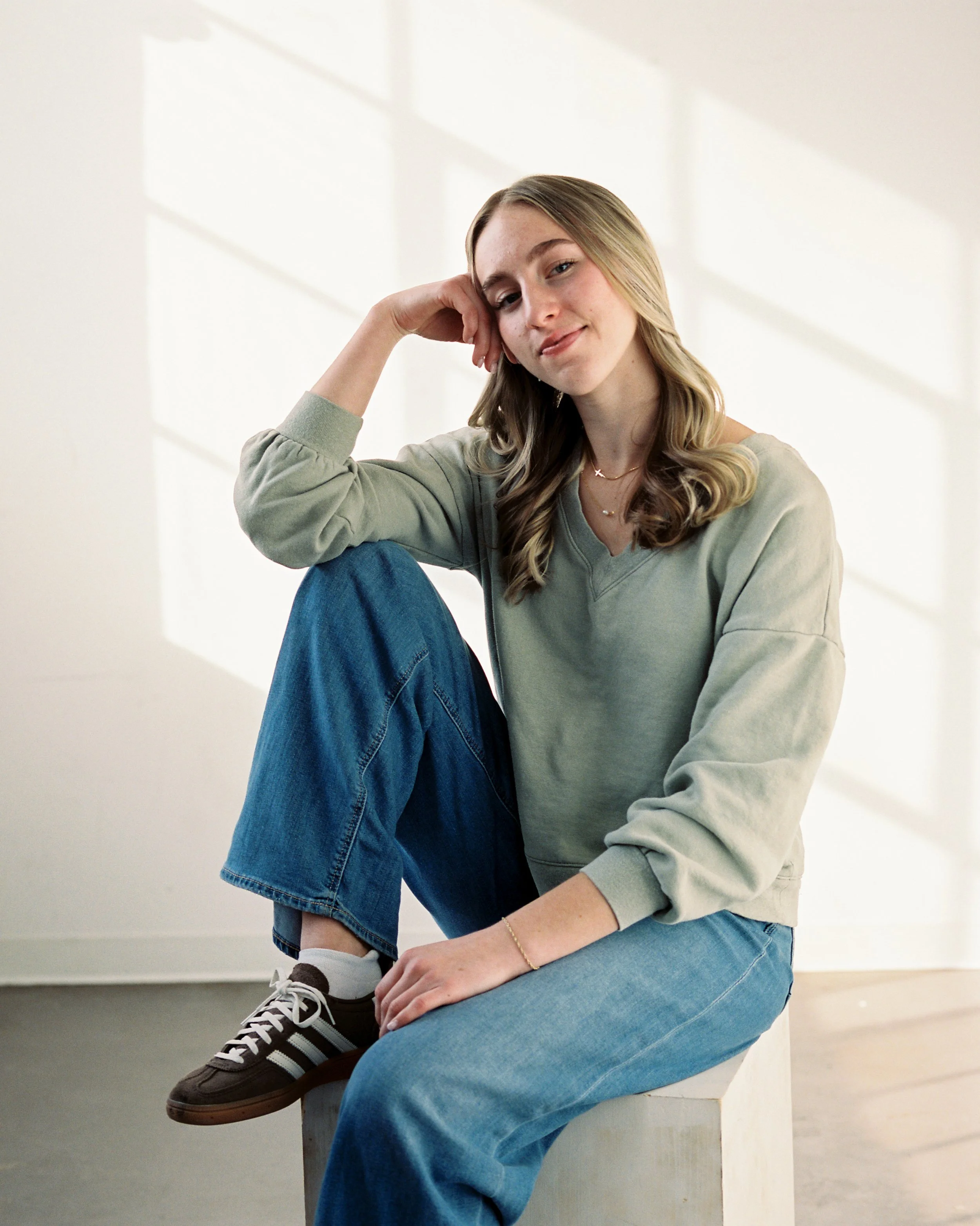 Portrait of a young woman with long blonde hair, wearing a light green sweatshirt and blue jeans, sitting on a white block in a brightly lit room with white walls and large windows, smiling slightly with her head resting on her hand.