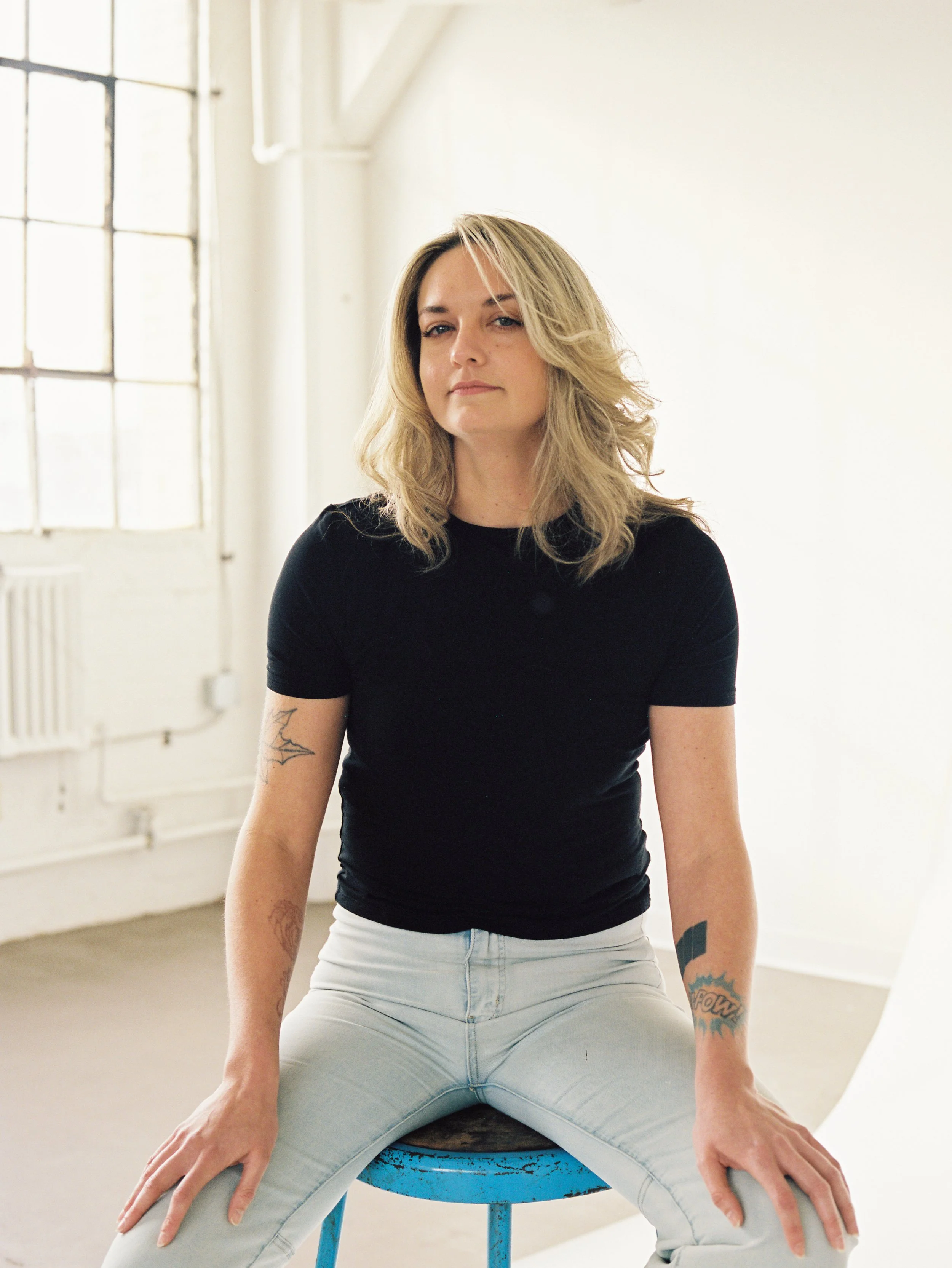 Portrait of a woman with blonde, wavy hair sitting on a blue stool in a bright room with large windows, wearing a black t-shirt and light-colored jeans, looking confidently at the camera.
