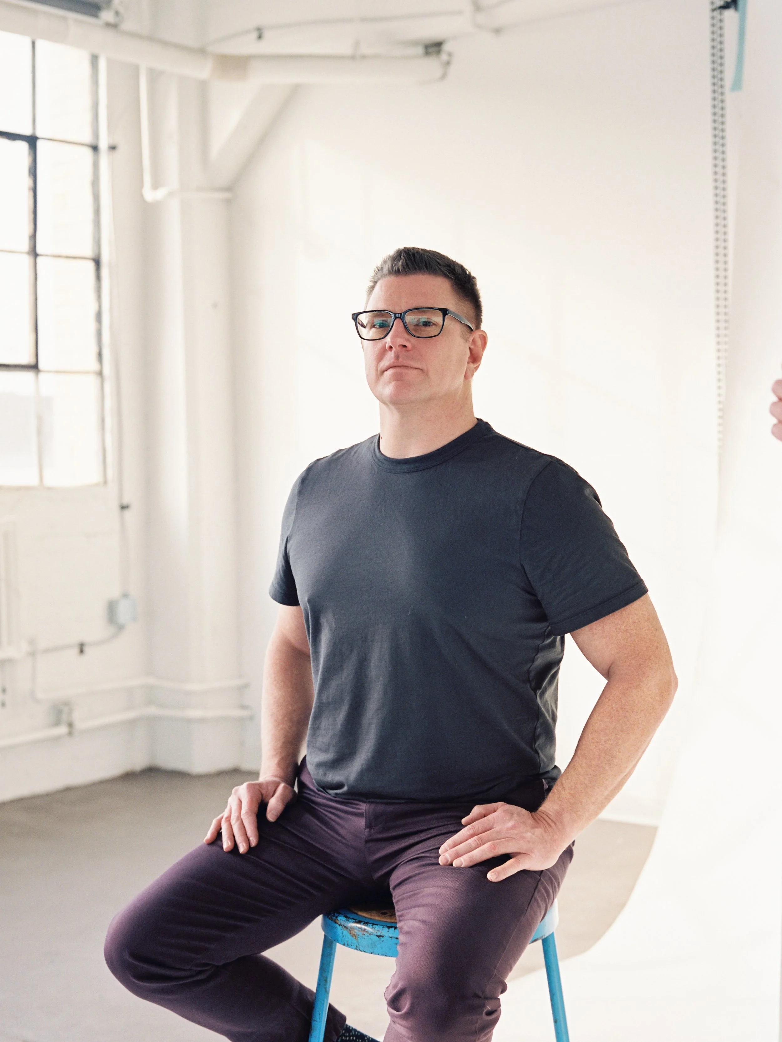 Portrait of a man wearing glasses and a black t-shirt sitting on a blue stool in a bright, minimalist studio with white walls and large windows.