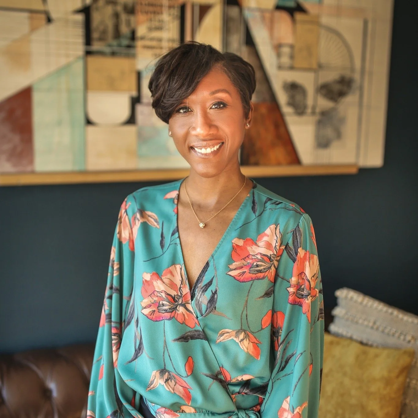 A woman with short dark hair and a warm smile, wearing a teal floral dress, standing indoors in front of a textured wall and a colorful abstract painting.
