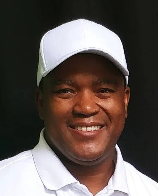 Smiling man wearing a white cap and white collared shirt against a dark background.