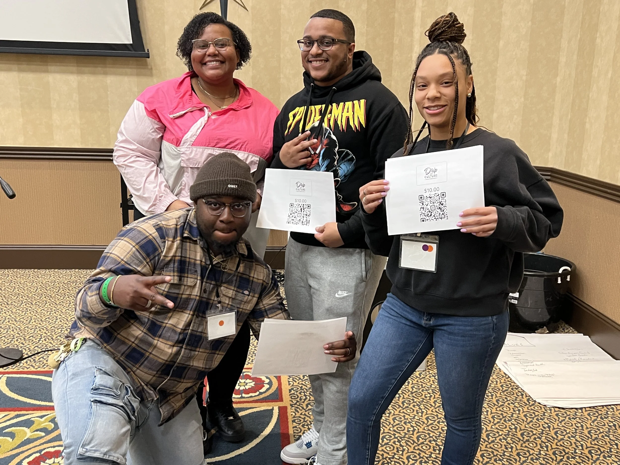 Group of five smiling adults posing indoors, holding printed certificates with QR codes and $10 value, with a patterned carpet and beige walls in the background.