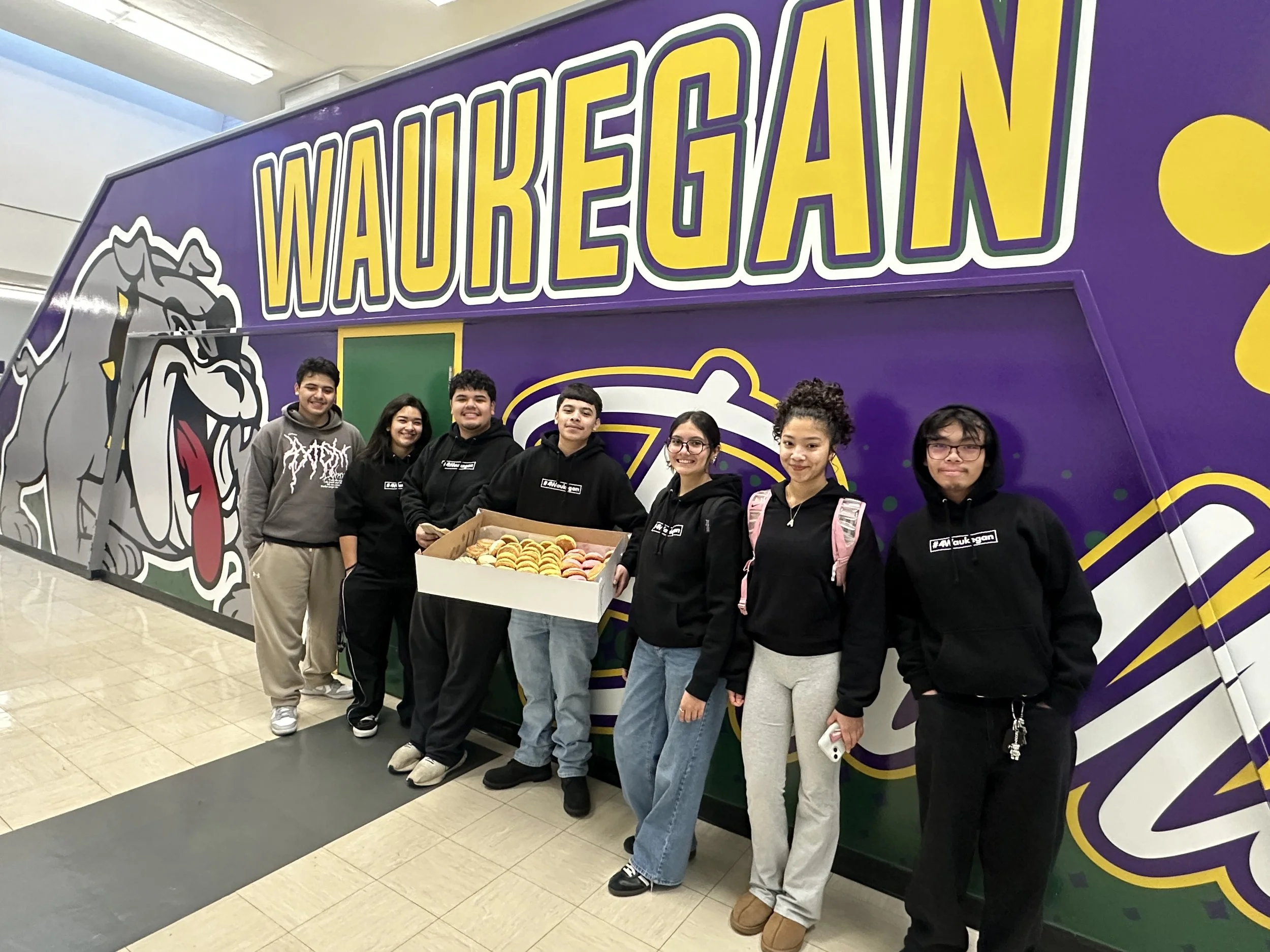 A group of seven teenagers standing in front of a large wall with the word 'WAUKEGAN' and a mascot logo.
