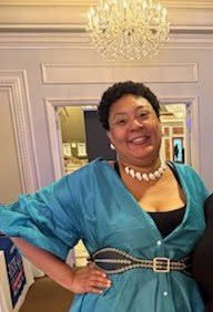 Woman smiling wearing a teal dress and pearl necklace, standing in an elegant room with a chandelier overhead.