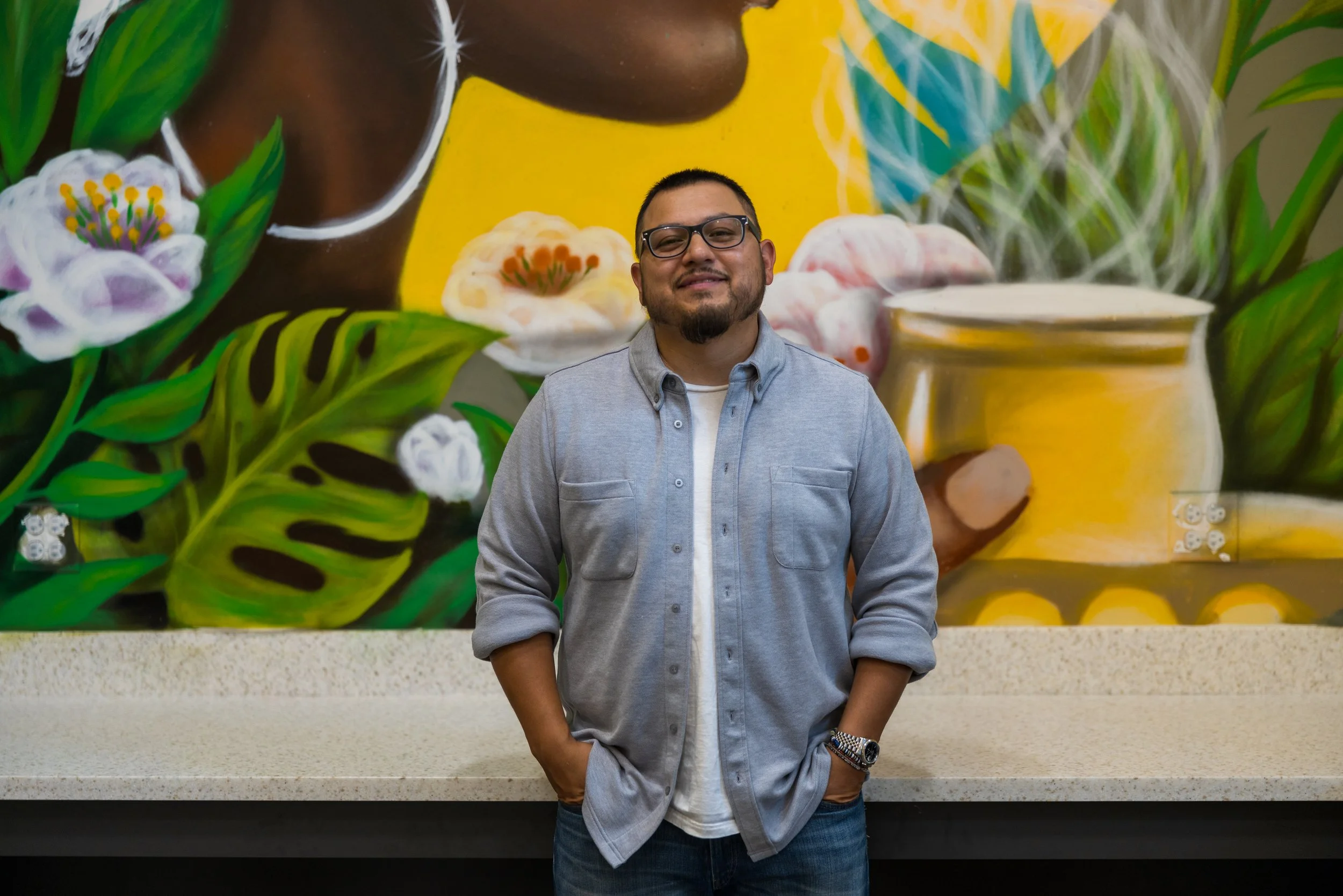 A man with glasses, a beard, and a goatee standing in front of a colorful mural, wearing a gray button-up shirt over a white t-shirt with hands in his pockets.