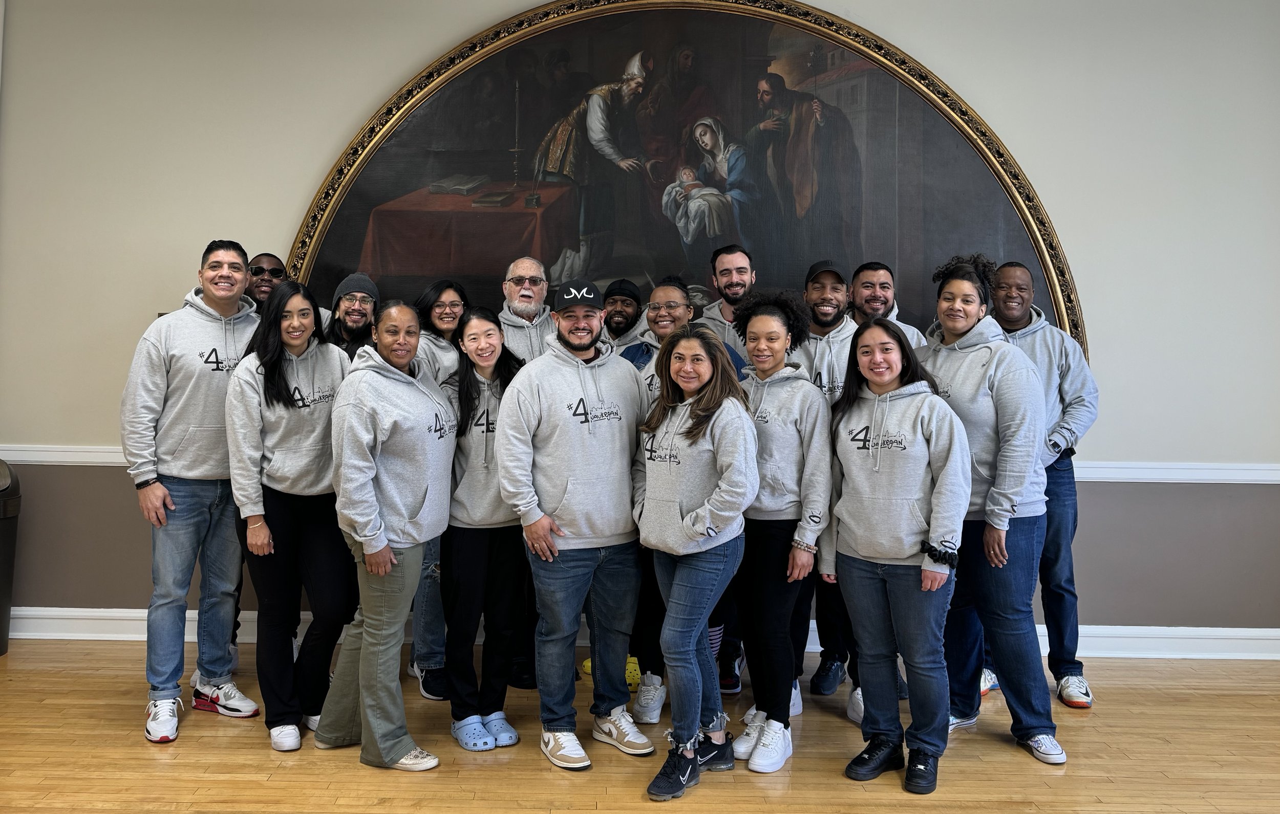 Group of smiling people wearing gray hoodies in front of a large religious painting on a wall, standing on a wooden floor.
