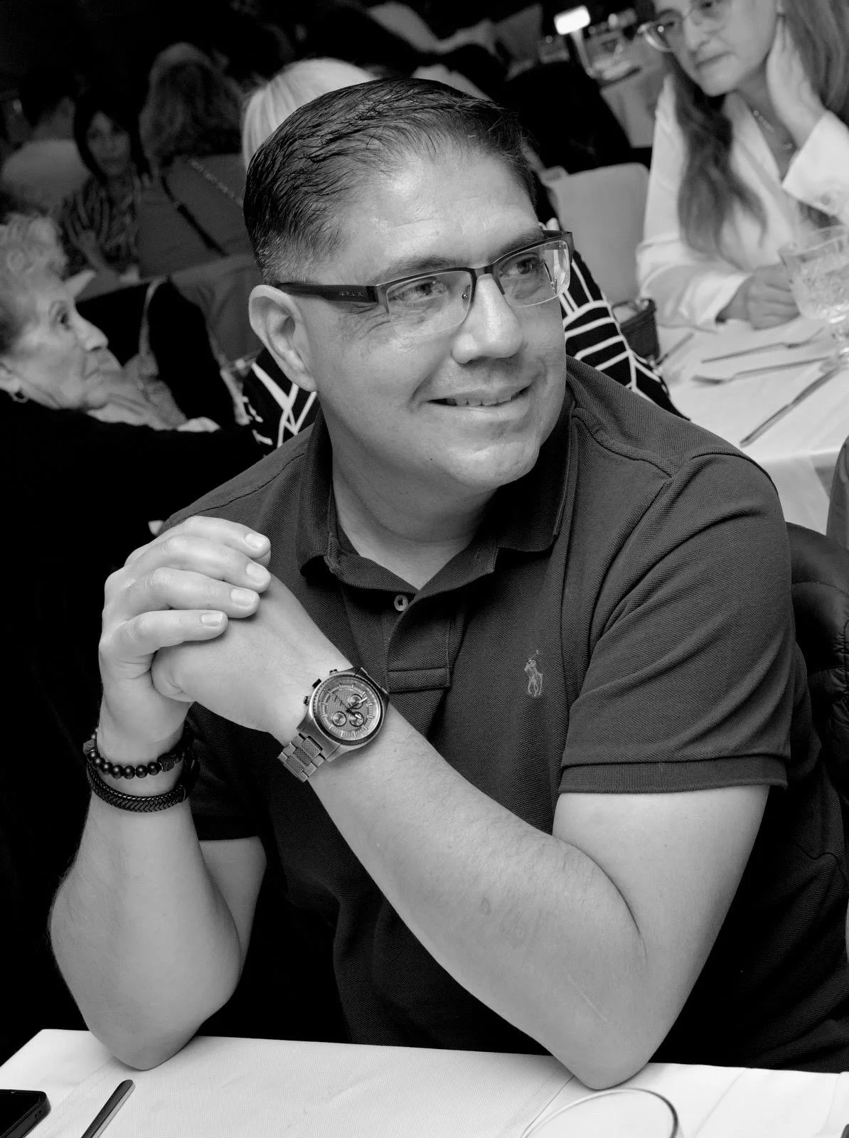 A man wearing glasses and a dark polo shirt, sitting at a table with his hands clasped, smiling, with other people in the background at a social gathering or restaurant.