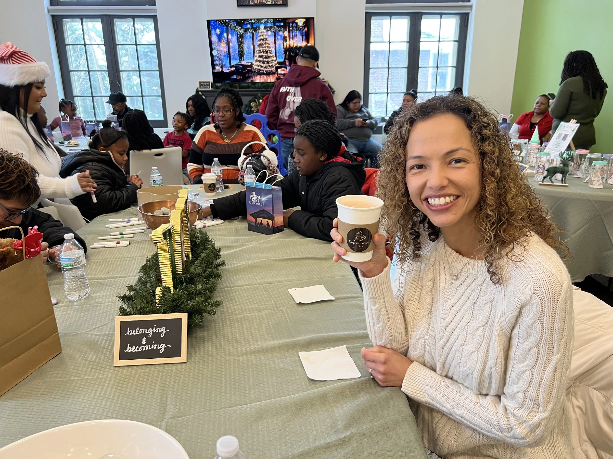 A woman with curly hair smiling and holding a cup of hot beverage at a festive gathering with children and adults seated at tables decorated with holiday ornaments and a small sign that says 'belonging & becoming'.