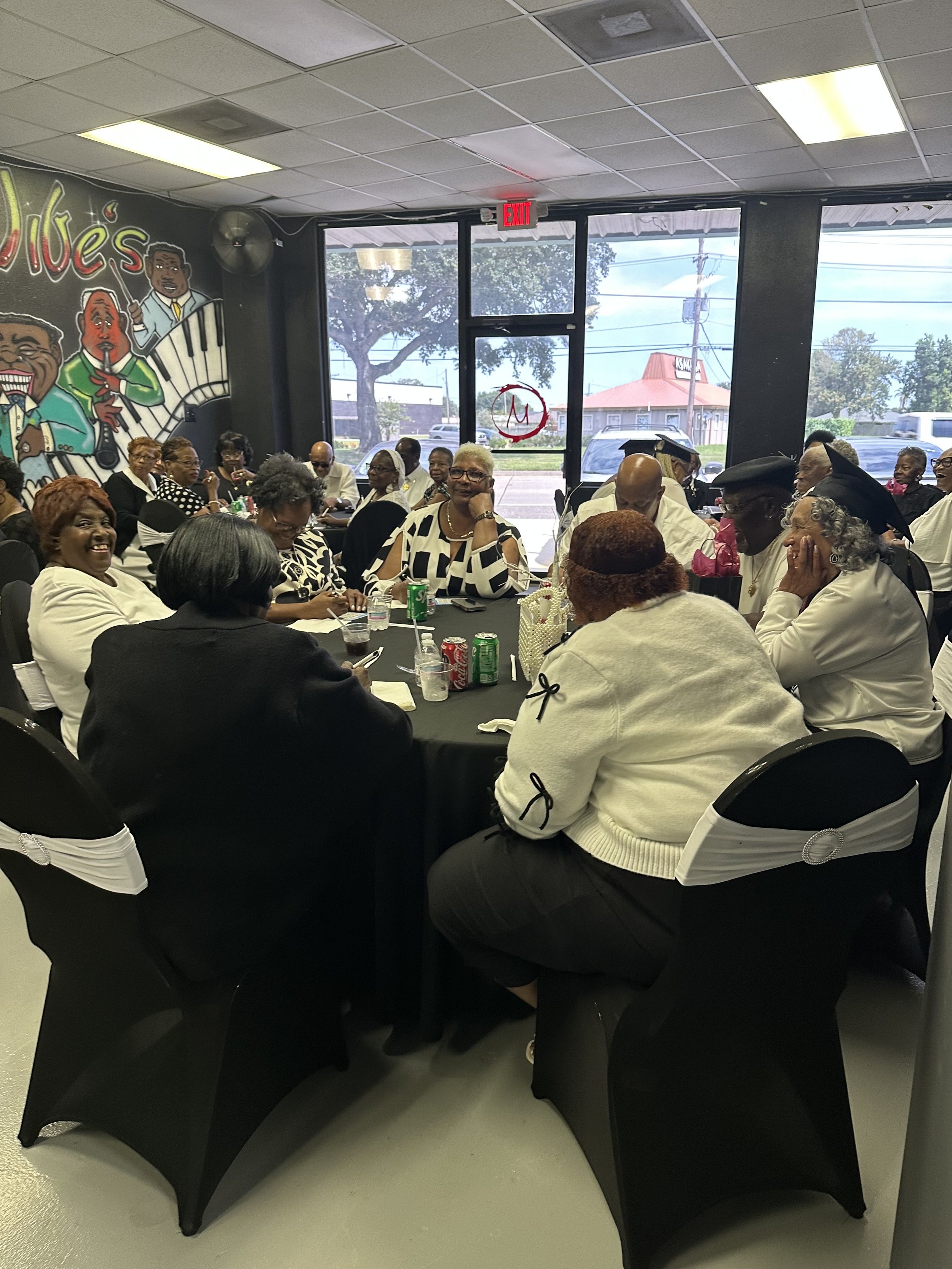 Group of adults sitting around a table in a restaurant, wearing black and white clothing, some with graduation caps, with a mural of jazz musicians on the wall