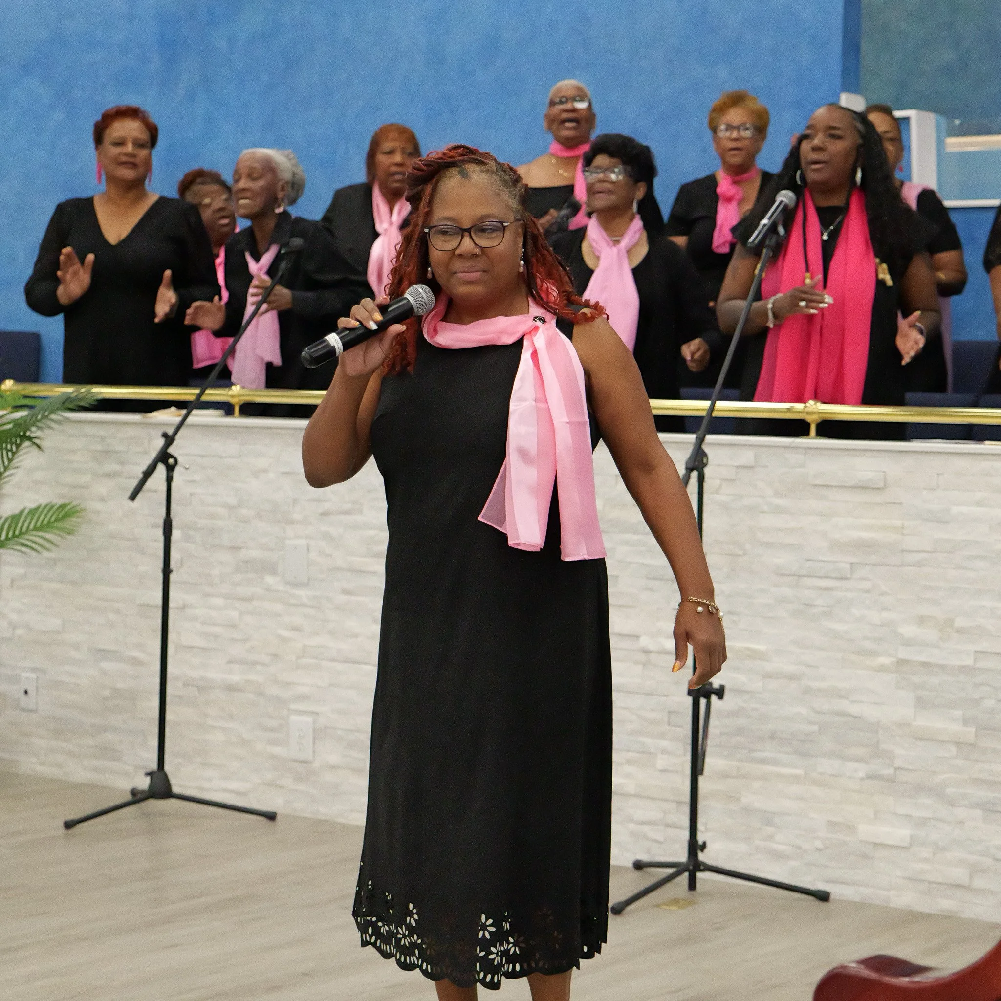 A woman in a black dress with lace trim and pink scarf holding a microphone, standing in front of a choir with black outfits and pink scarves, inside a church or auditorium.