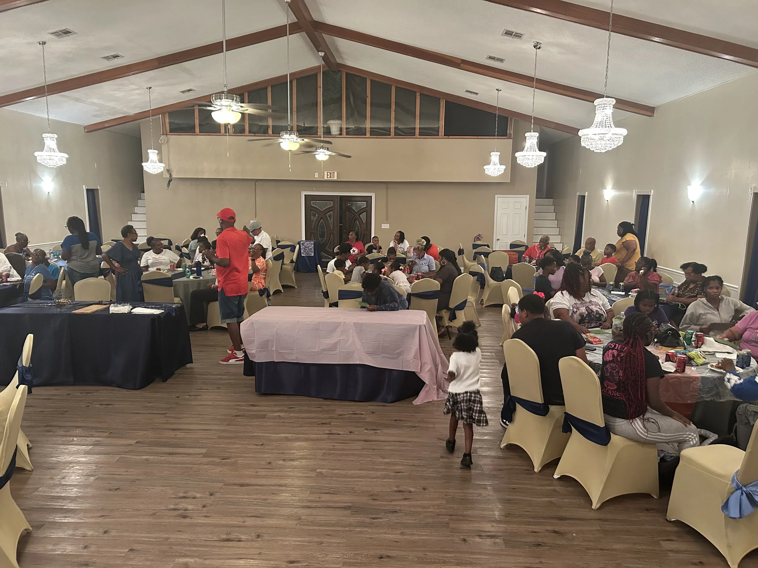 People gathered in a decorated banquet hall with round tables, chairs with blue sashes, and chandeliers, attending a social event or celebration.