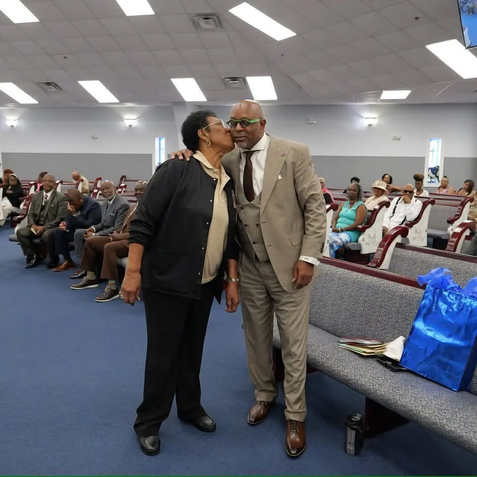 A woman kisses a man on the cheek inside a church, with several people seated in the background.