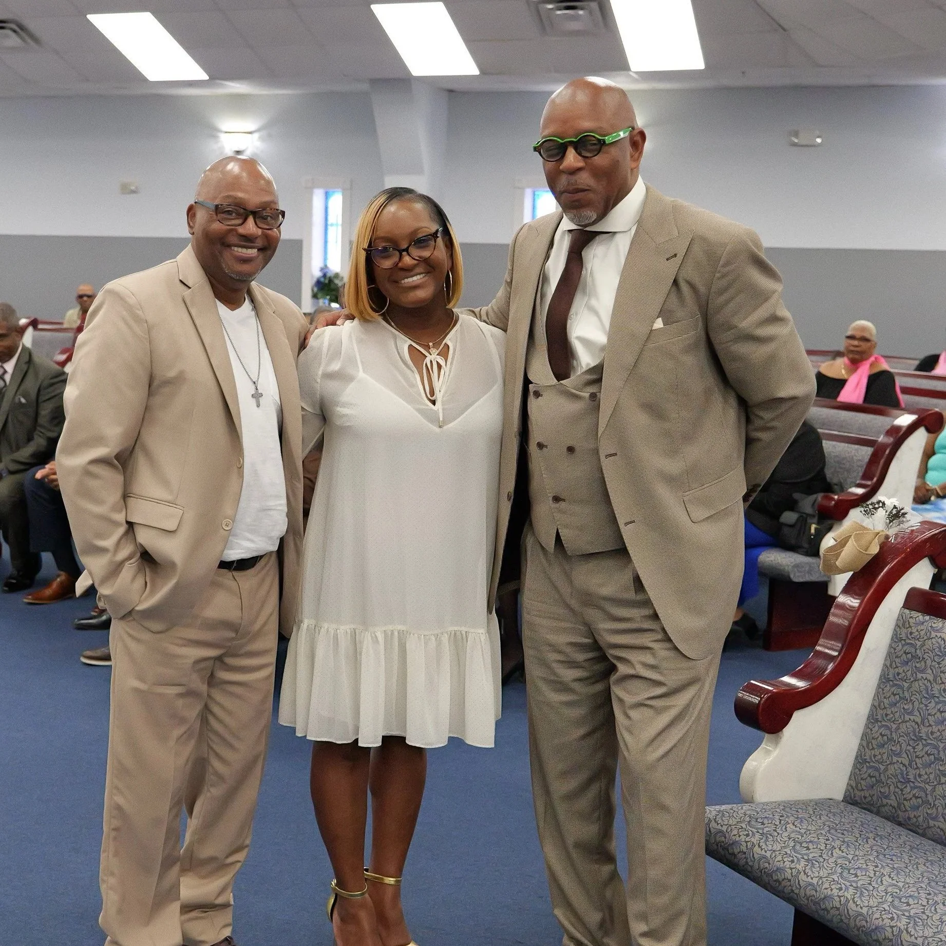 Three adults standing together inside a church, smiling at the camera. Two men wear beige suits, and a woman in the middle wears a white dress. In the background, other people are seated in pews.