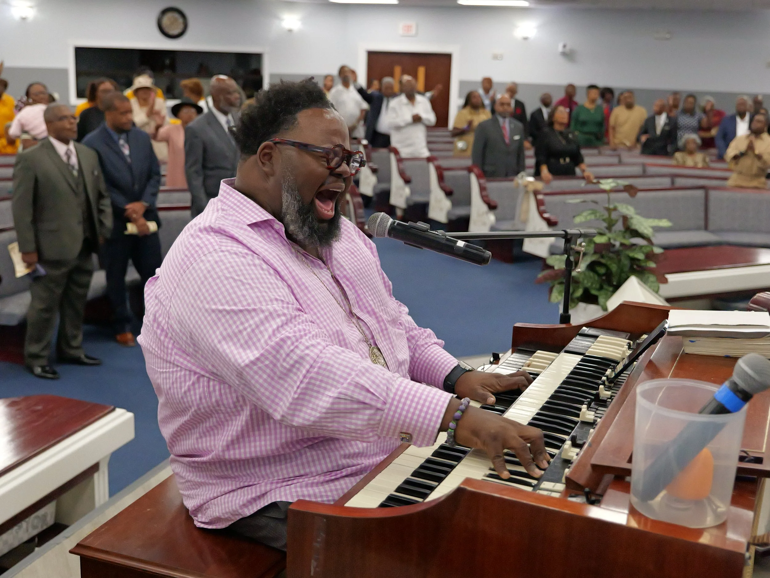 A man in a pink checkered shirt playing a keyboard and singing into a microphone at a church service, with a congregation in the background.