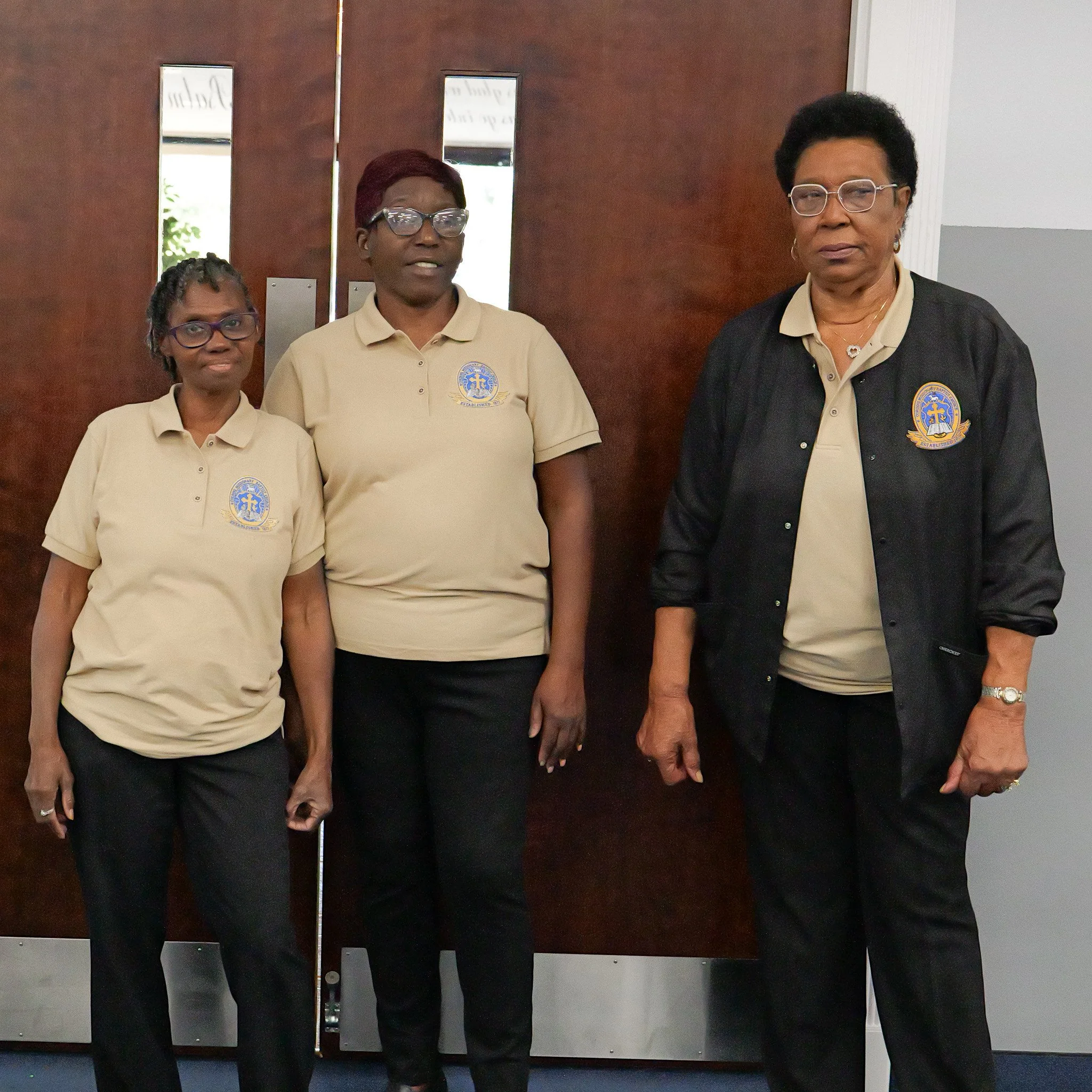 Three women standing indoors in front of a wooden door, wearing matching beige polo shirts with a logo, and black pants. The woman on the right wears a black jacket over her polo shirt.