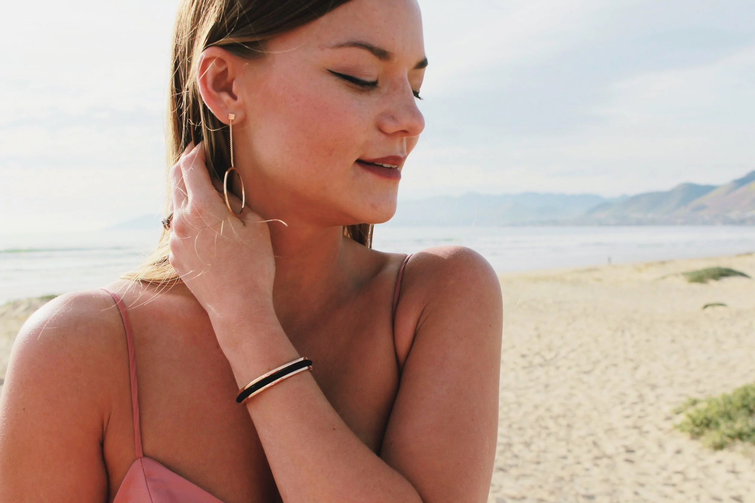 Gold Dangle Earrings with Pink Satin Dress and Gold Floral HairTie Bracelet with a beach Background. 