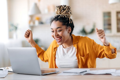  Overjoyed excited african american girl with dreadlocks, freelancer, manager working remotely at home using laptop, looks at screen with surprise, smiling face, gesturing with hands, got a dream job 