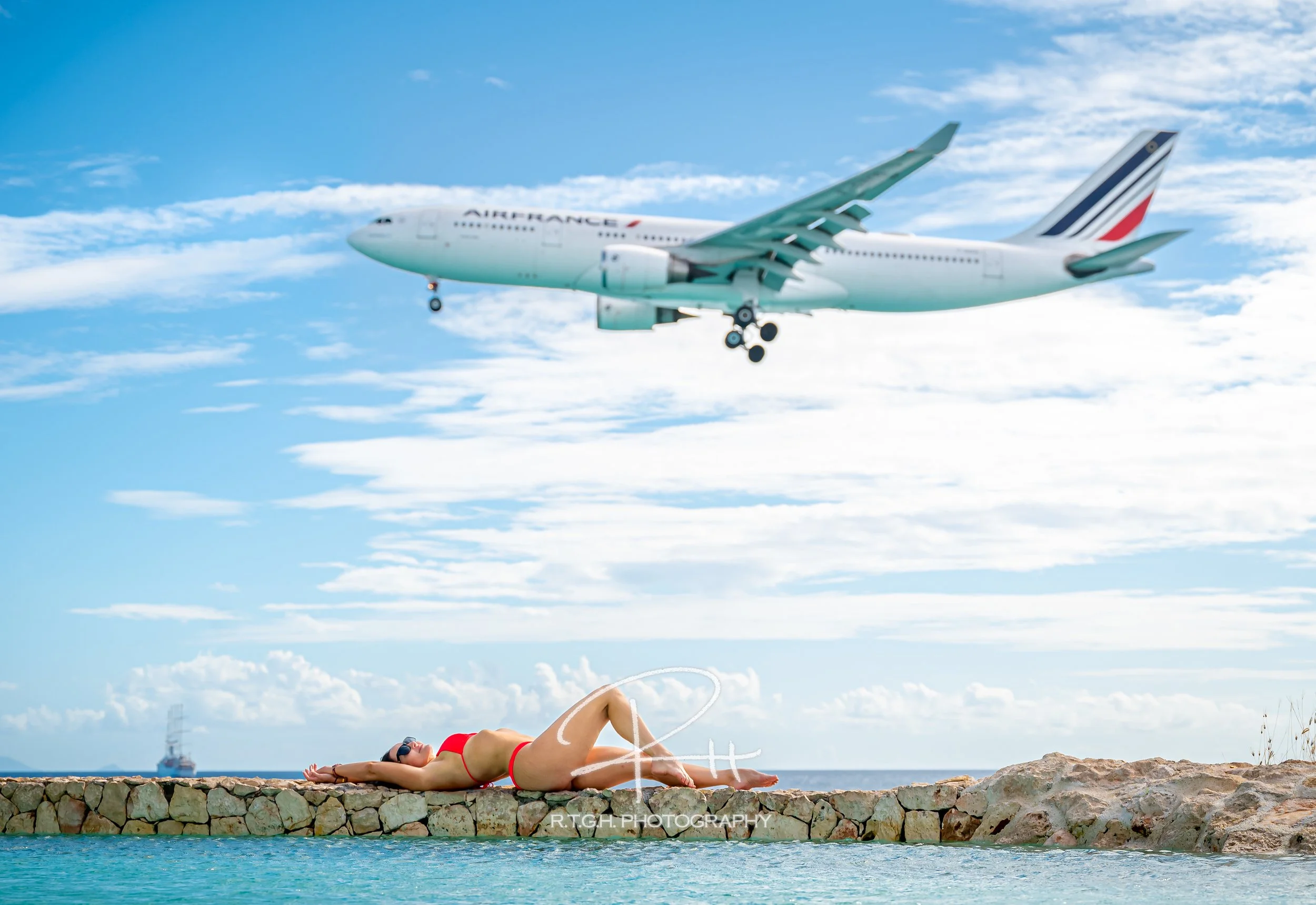 A woman in a red swimsuit and sunglasses lying on a stone platform near water, with an airplane flying in the sky above.