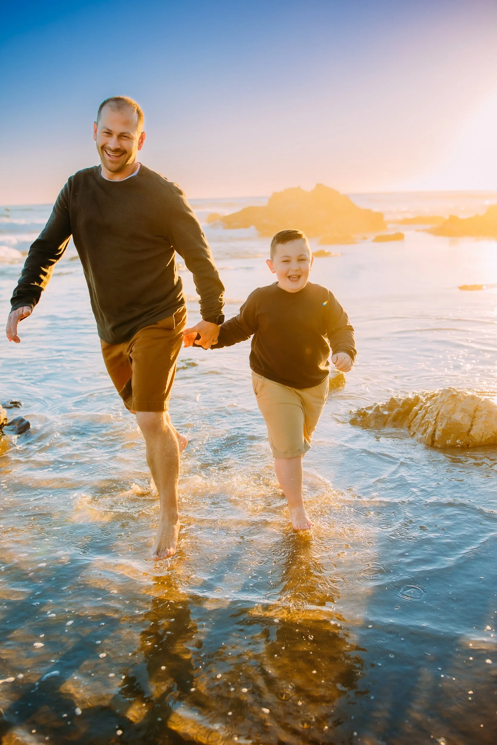 dad and son holding hands and laughing and running in the ocean tide during their family photos in Newport Beach.
