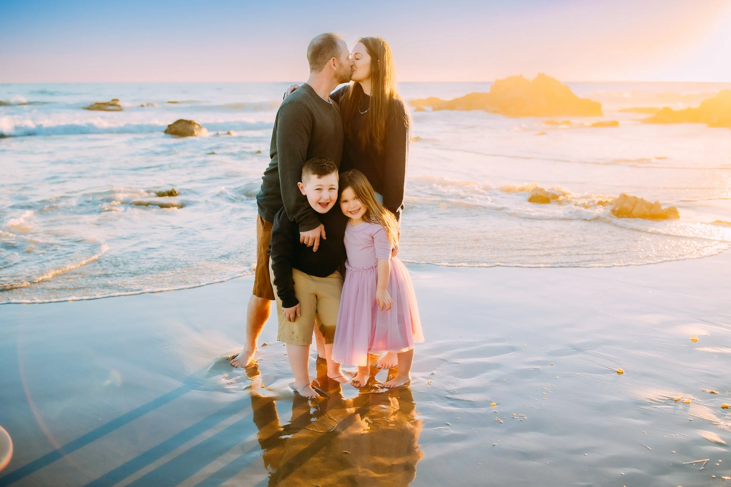 siblings laughing at the camera while their mom and dad kiss at sunset during their family photo session in Newport Beach.