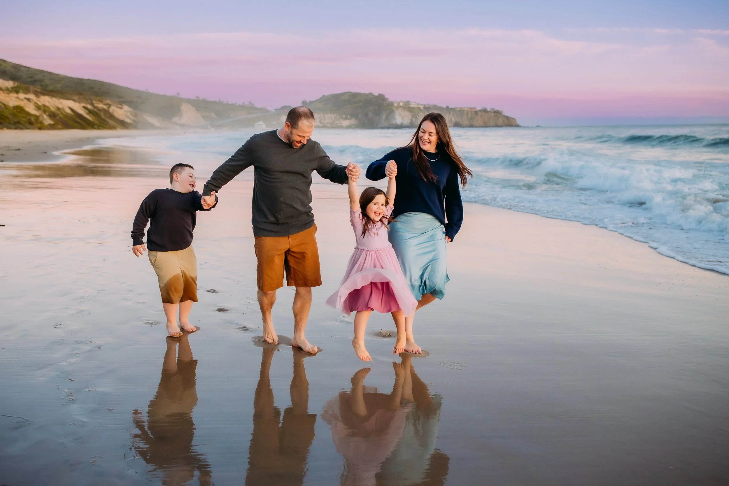 family laughing and swinging daughter into the air during their ocean photo session in Newport Beach.