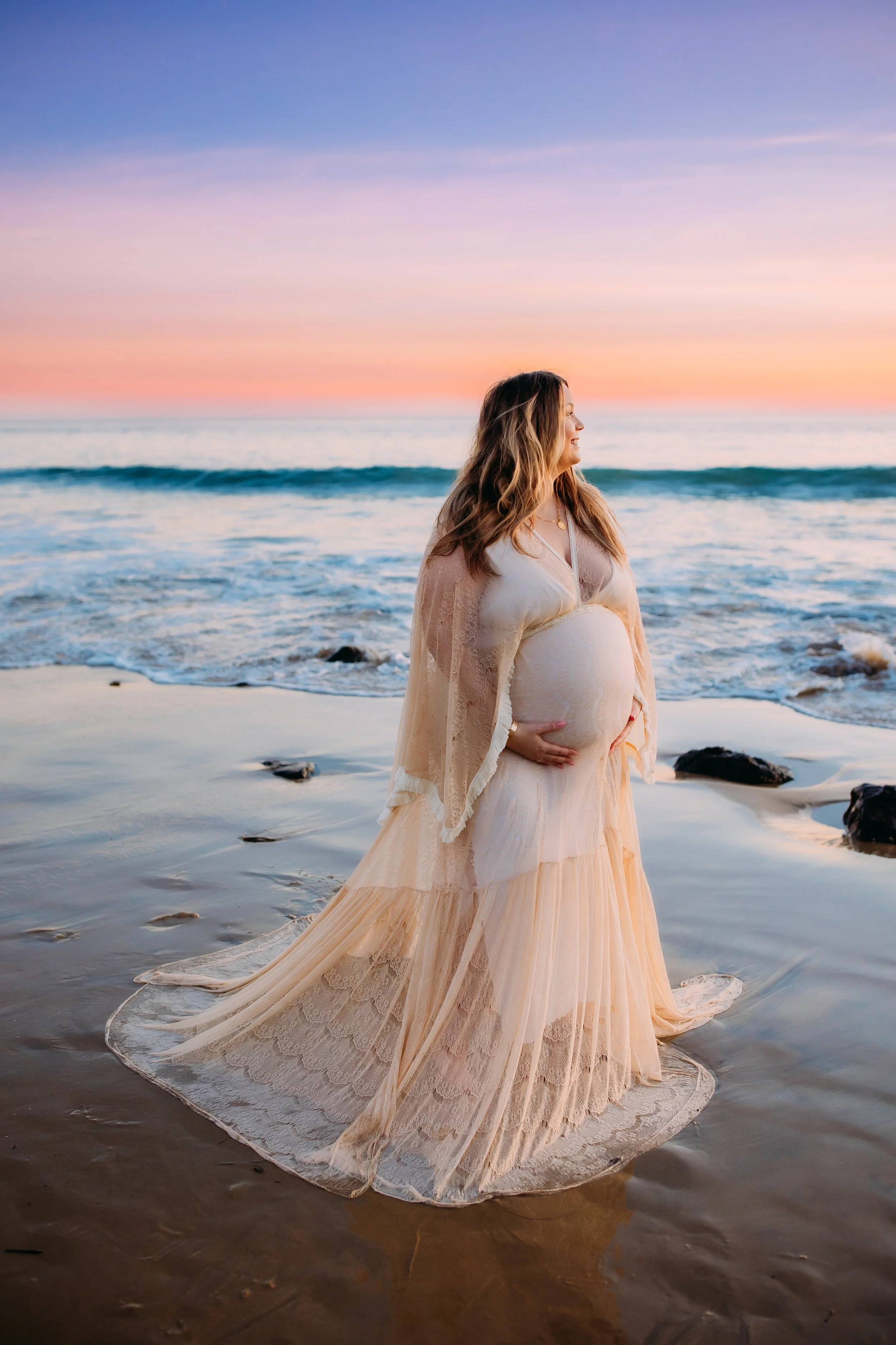 woman holding her pregnant belly as she smiles off into the sunset during her beach photoshoot in Newport Beach, Orange County, California