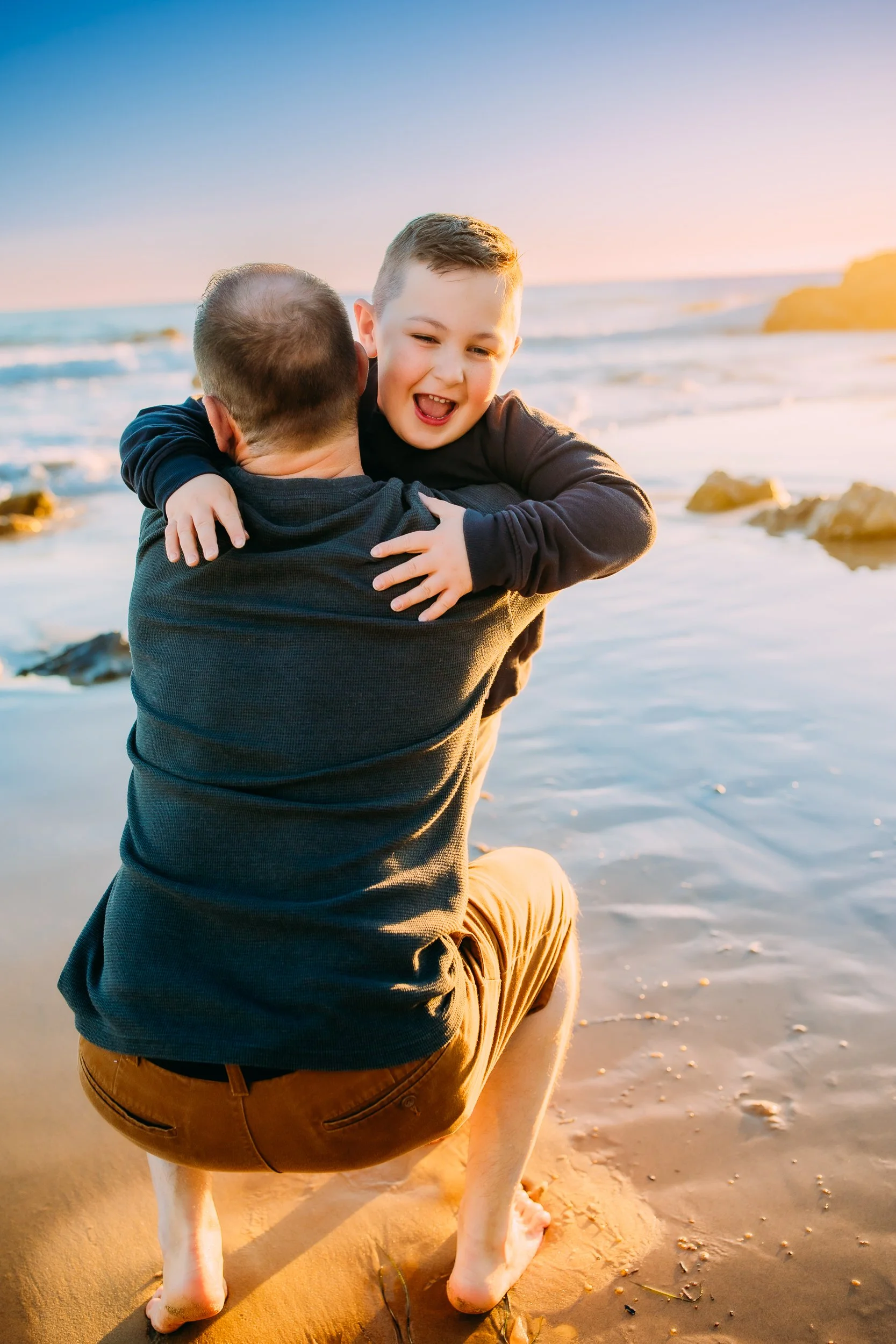 son running to his dad for a large embrace during their family photoshoot at the ocean in Newport Beach.