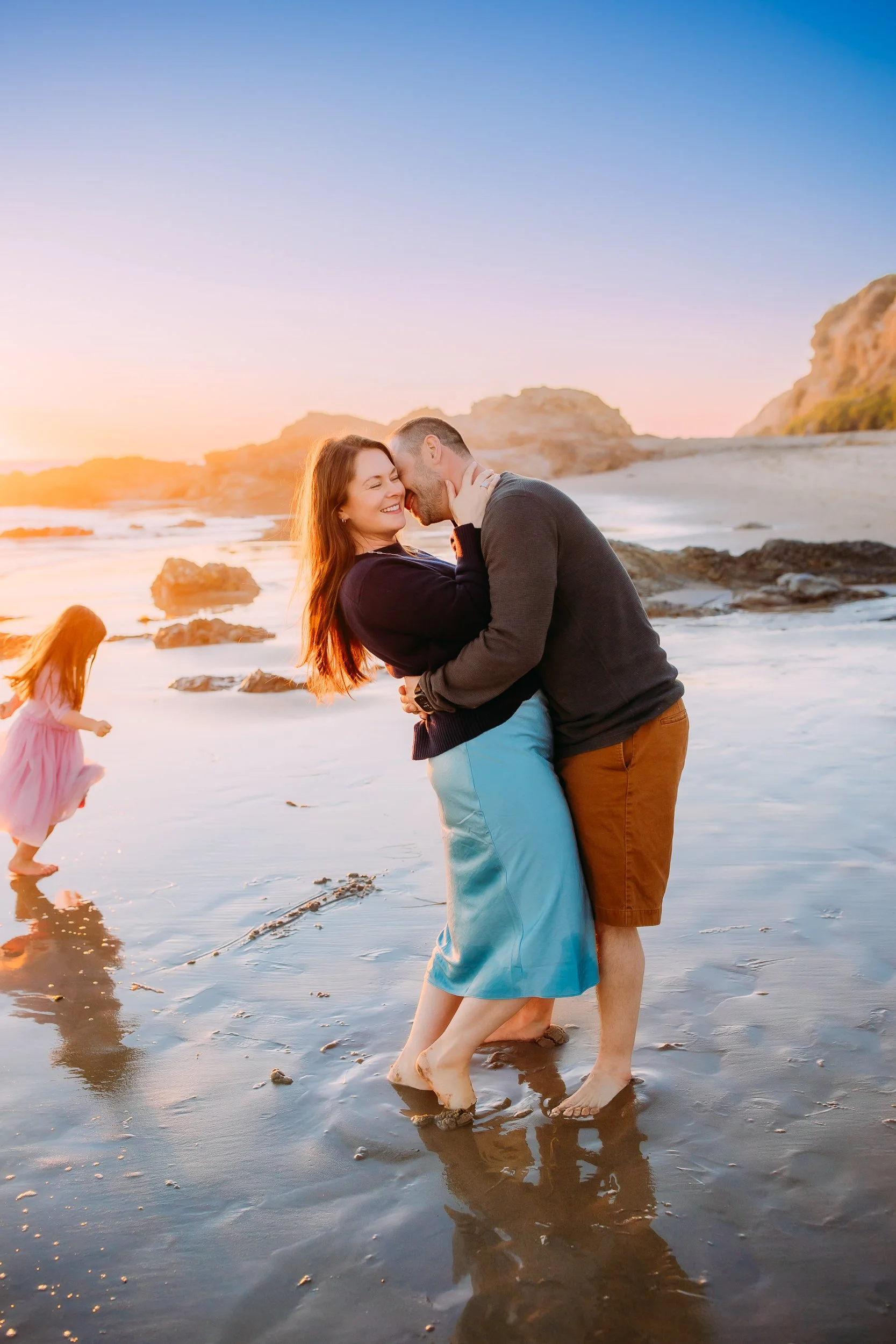 husband and wife laughing and embracing while their daughter runs in the background behind them during their sunset family photo session in Newport Beach.