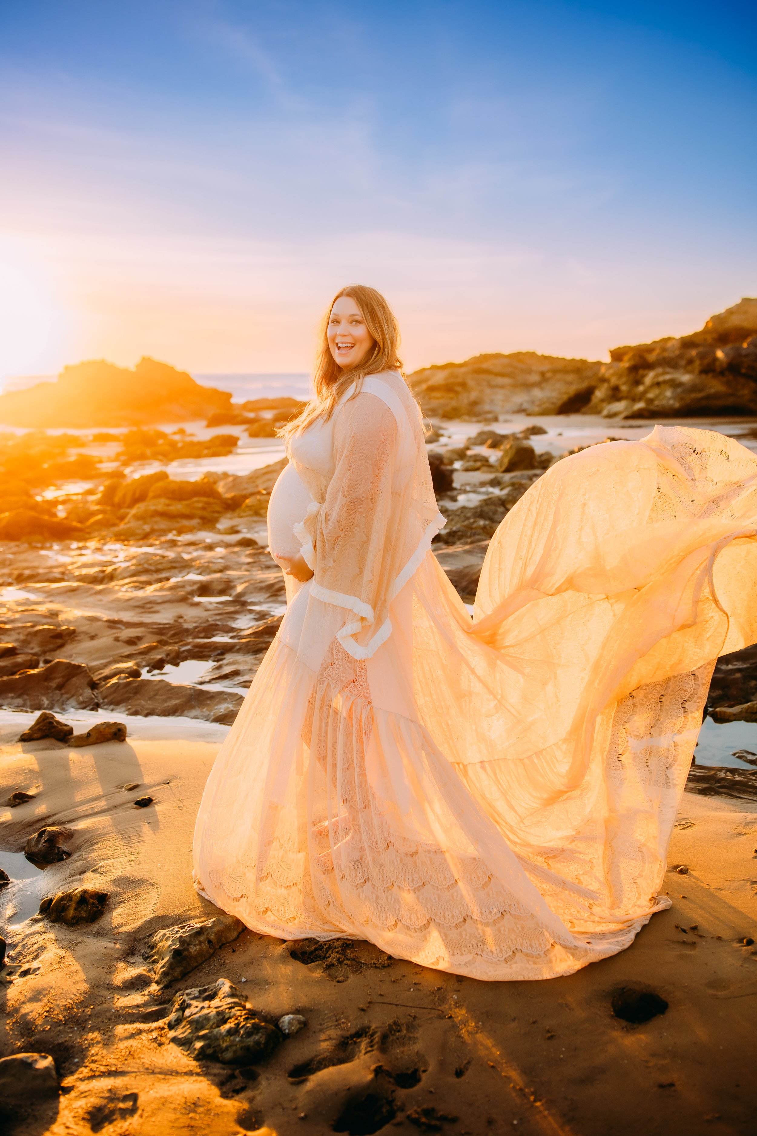 mother to be holding her belly and smiling into the camera during her photo session at sunset in Newport Beach, California