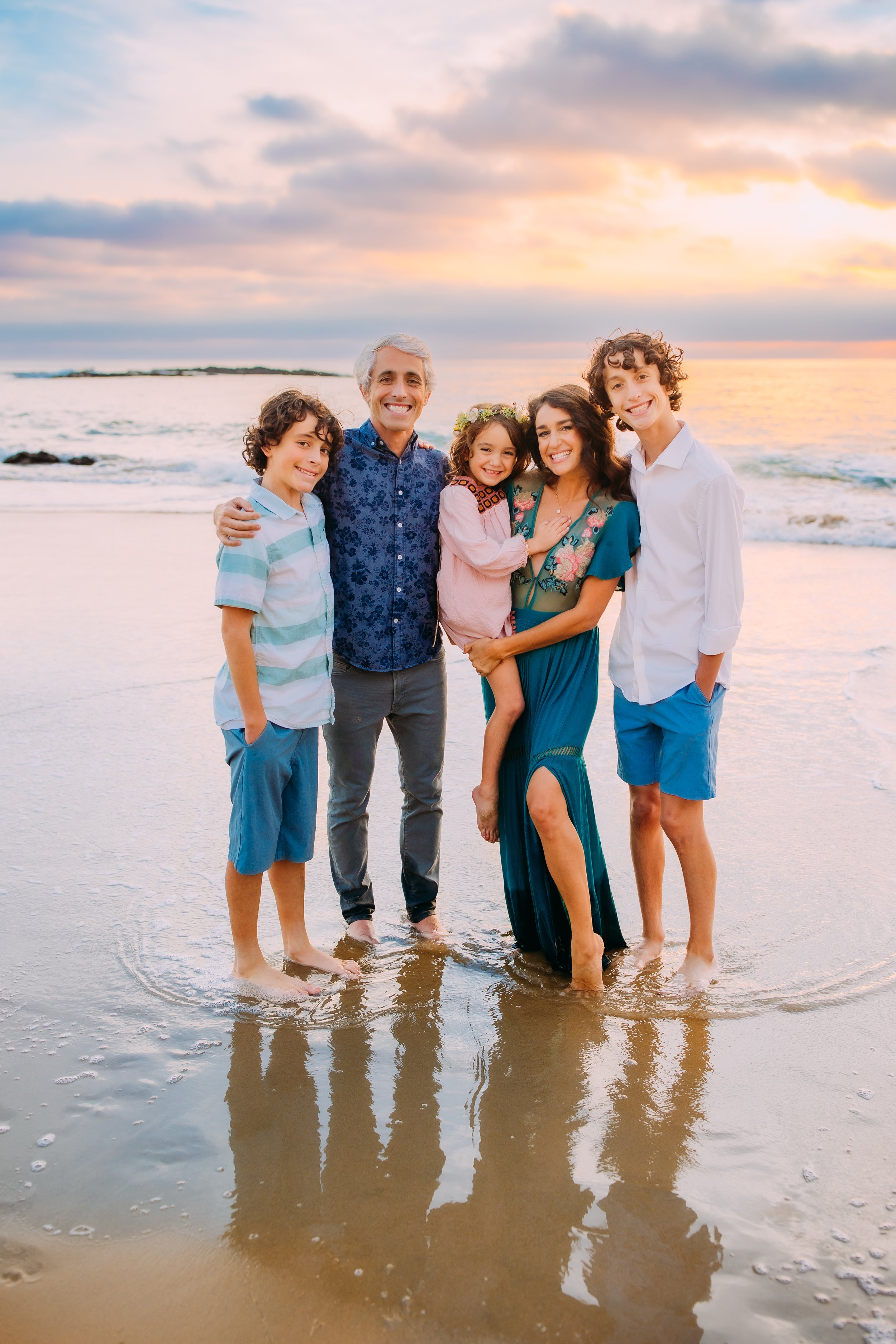 family of five posing for the camera while standing in the tide water at the ocean at sunset